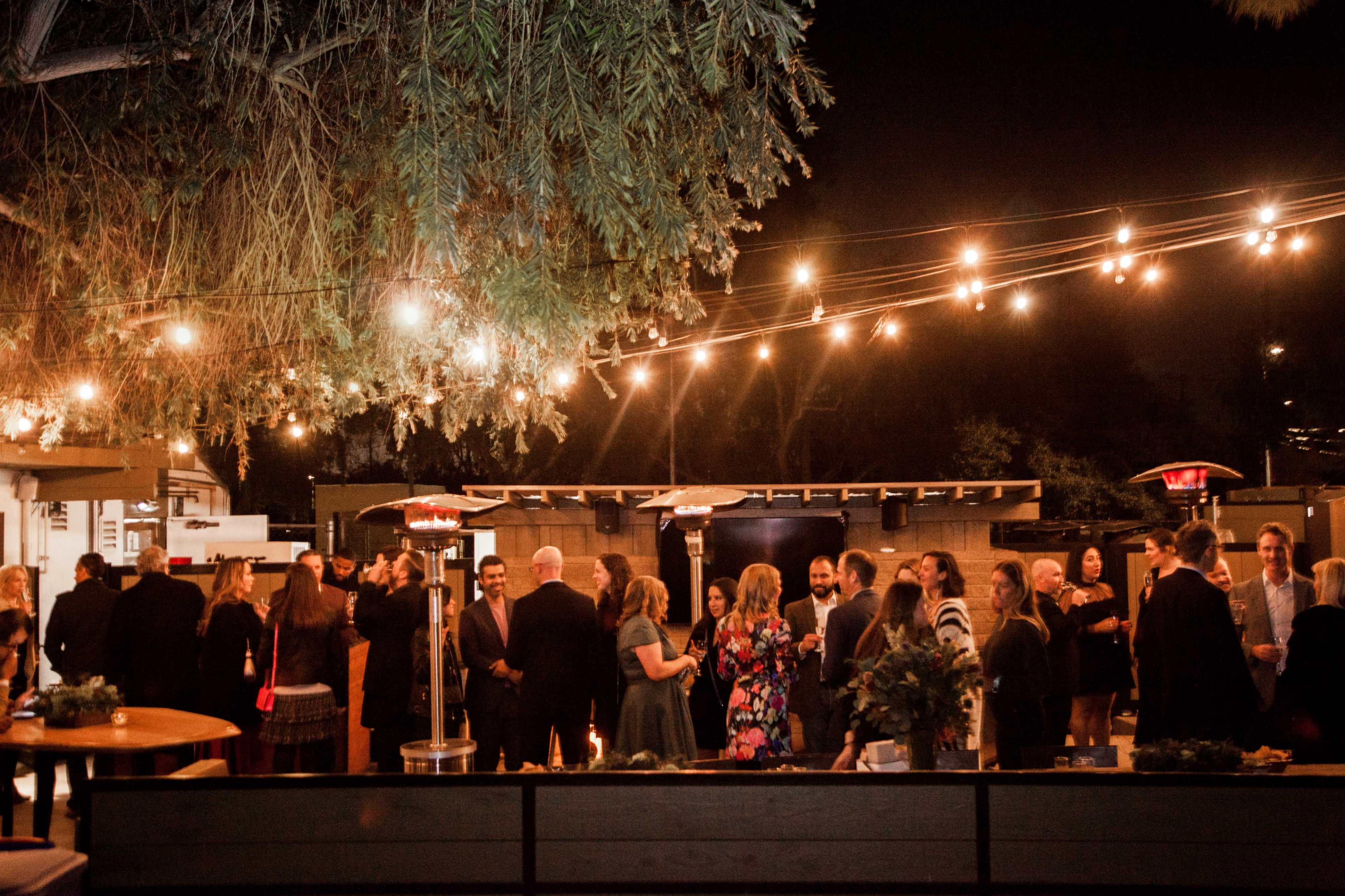 A gathering of people socializing at an outdoor venue, illuminated by string lights and heat lamps, during the evening.