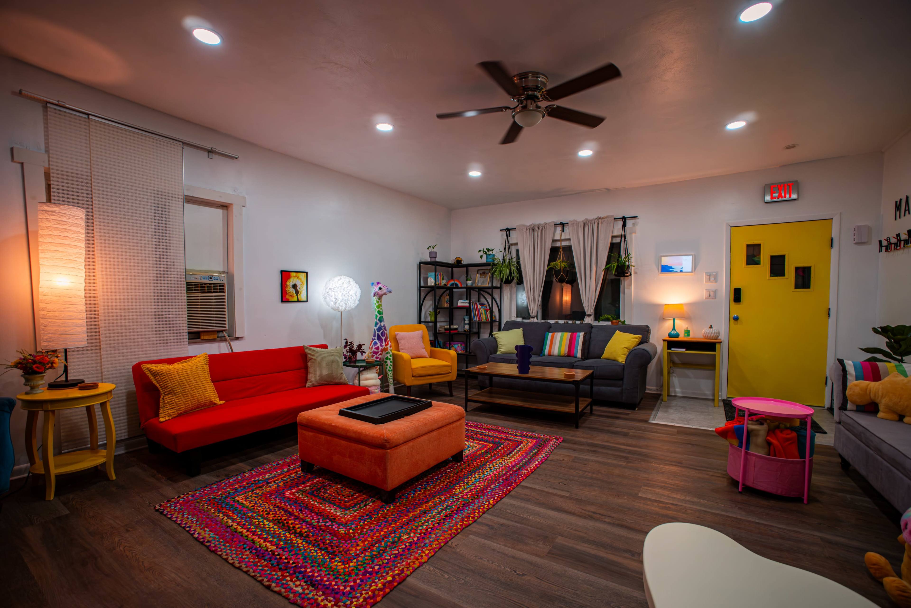 The image shows a brightly colored living room with a mix of seating options, a patterned rug, and a vibrant yellow door.