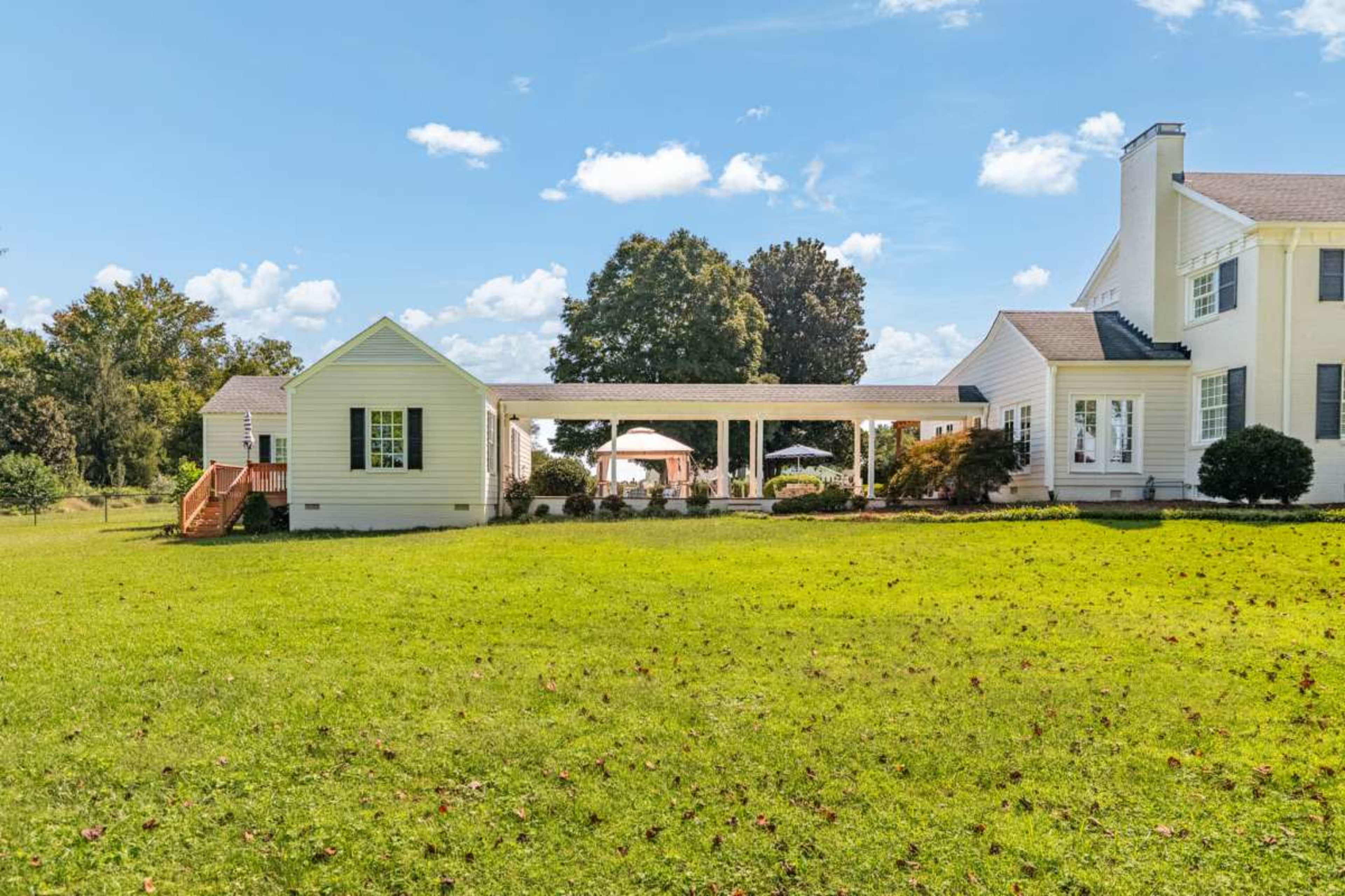 A large white house with a porch connects two sections, surrounded by a grassy lawn and a blue sky with fluffy clouds.