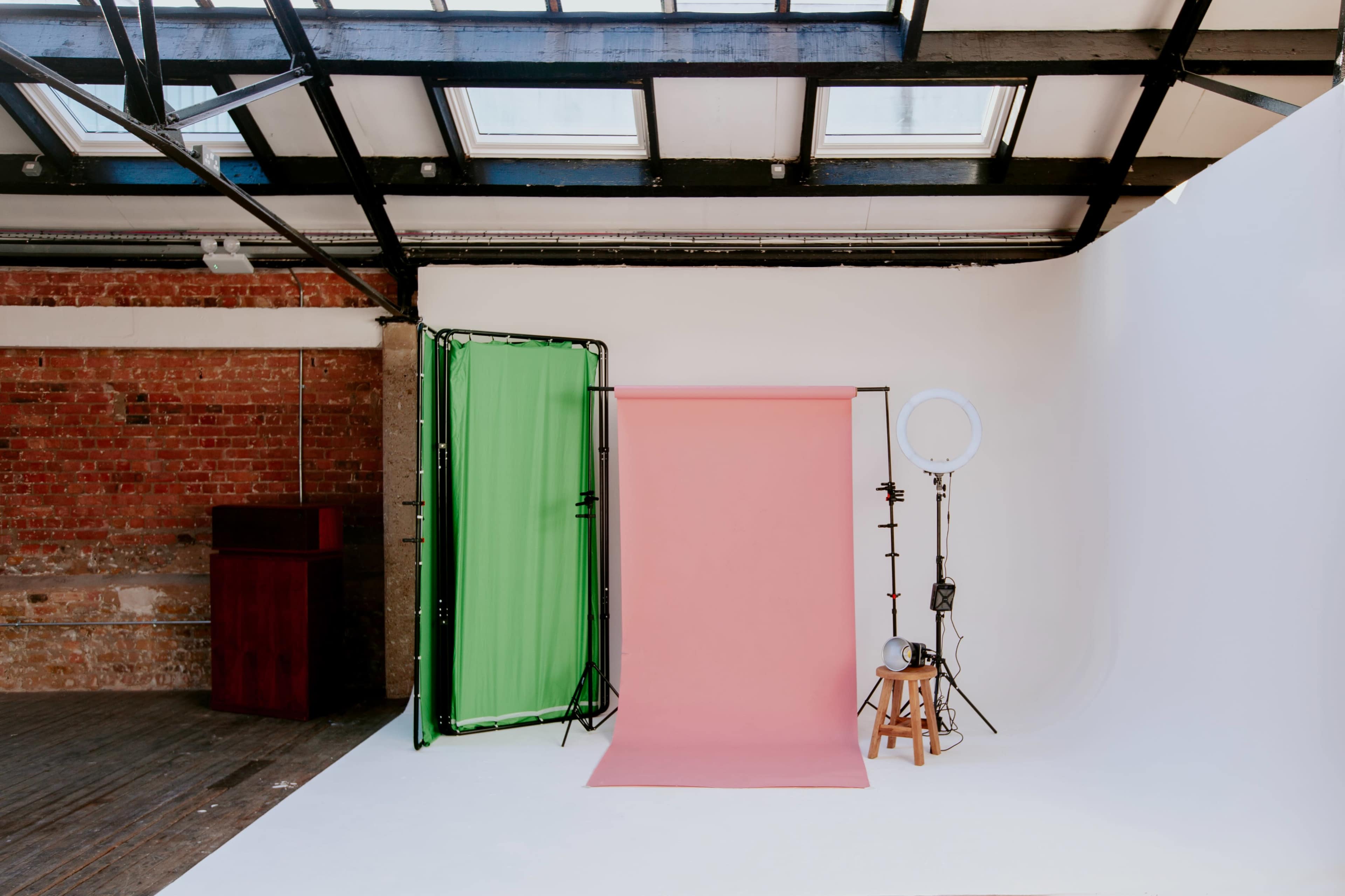 The image shows a photography studio with a pink backdrop, a green screen, lighting equipment, and a wooden stool.