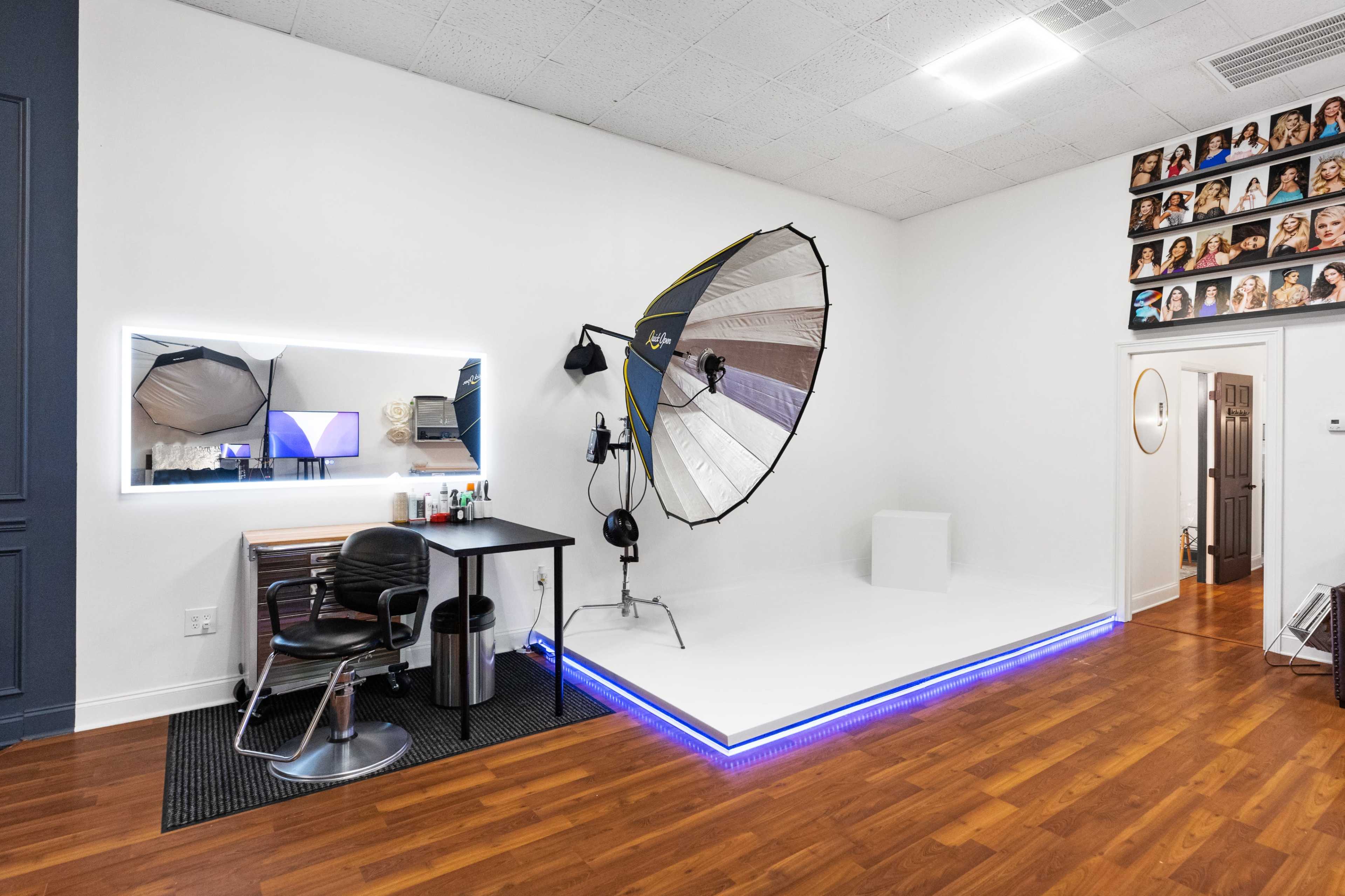The image shows a photography studio featuring a lighting umbrella, an elevated white platform, a vanity desk with a chair, and a wall adorned with framed portraits.