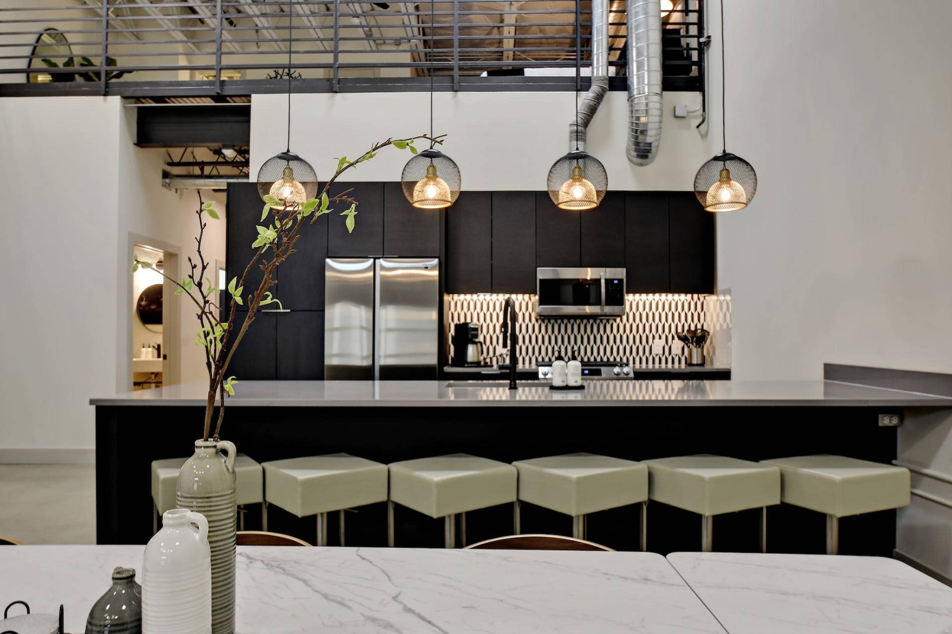 A modern kitchen with sleek black cabinetry, stainless steel appliances, and a marble dining table with a view of a bar area featuring light-colored stools.