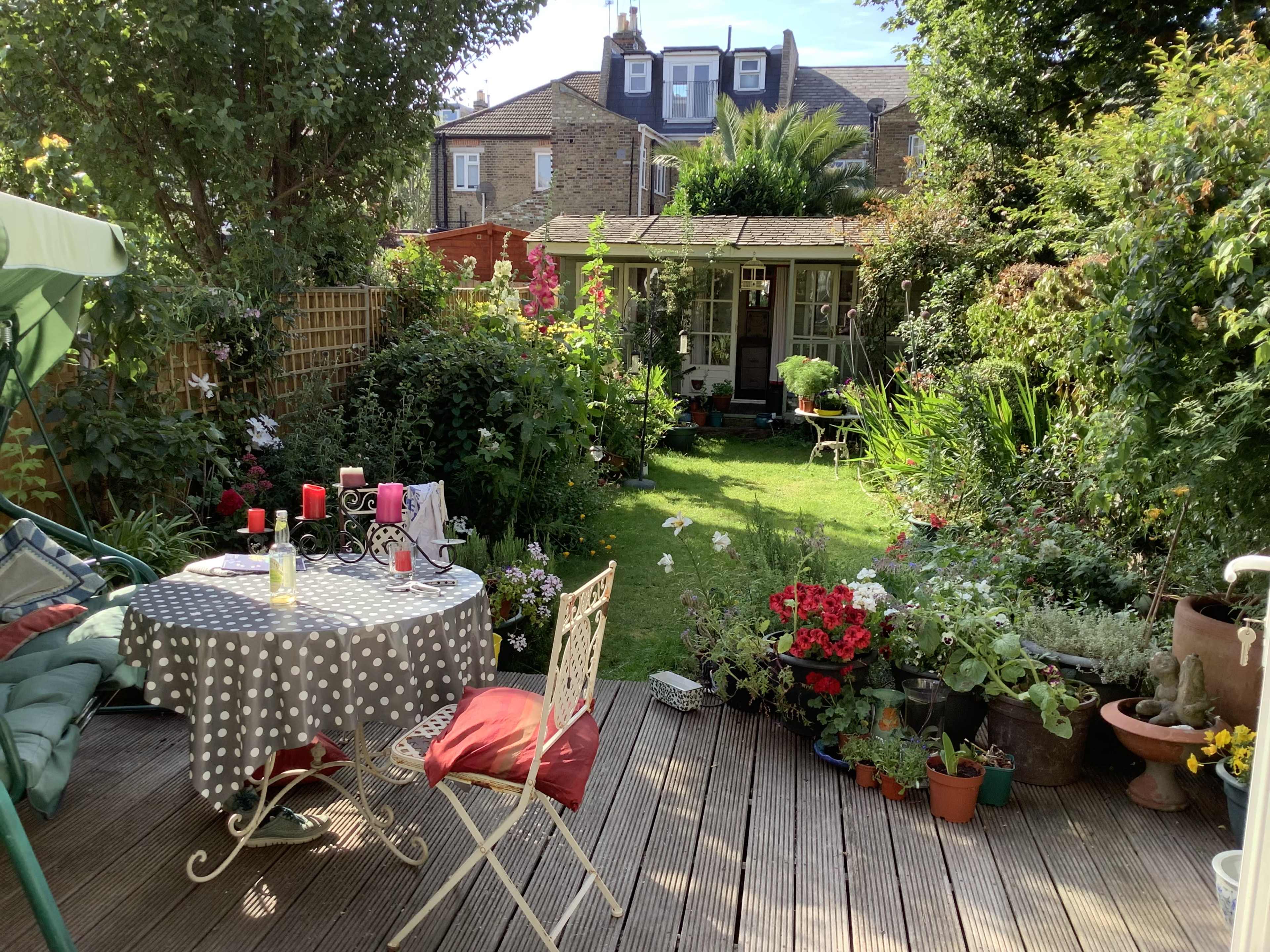 A wooden deck features a polka-dot table set for dining, surrounded by a lush garden filled with various plants and flowers, leading to a small wooden shed.