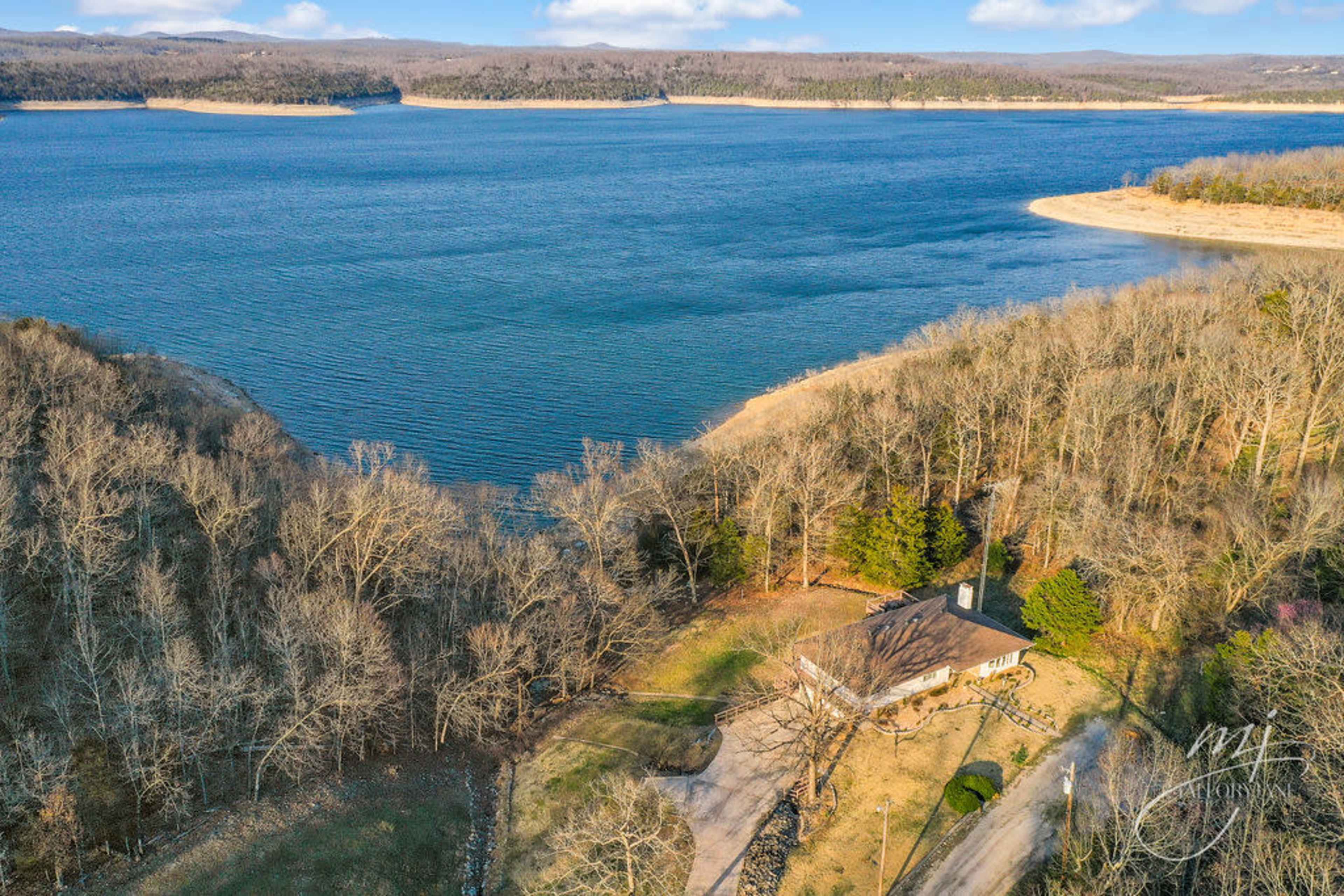The image shows a house situated near the shore of a large lake, surrounded by trees and hills in the background.