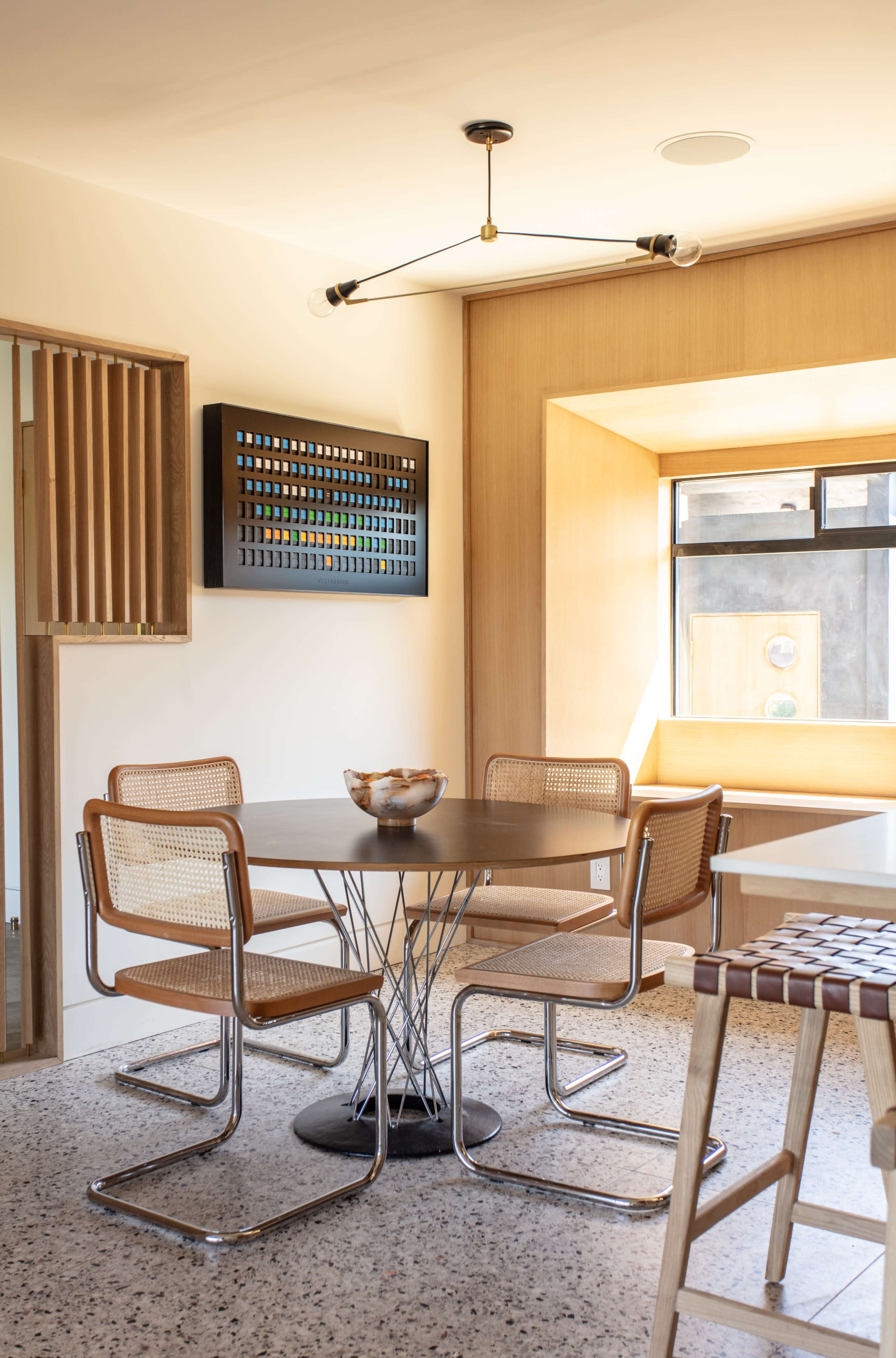 A modern dining area features a round table surrounded by four chairs, with a wall-mounted artwork and a window letting in natural light.