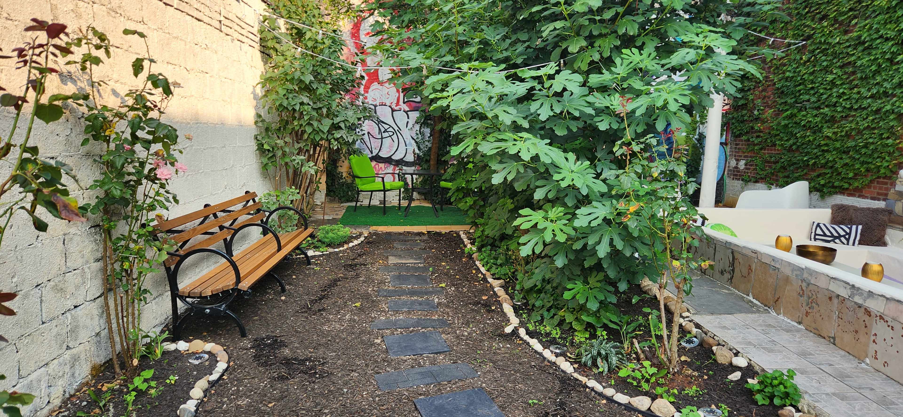 The image shows a small garden with a stone path, a wooden bench, and lush greenery, bordered by a wall and a seating area.