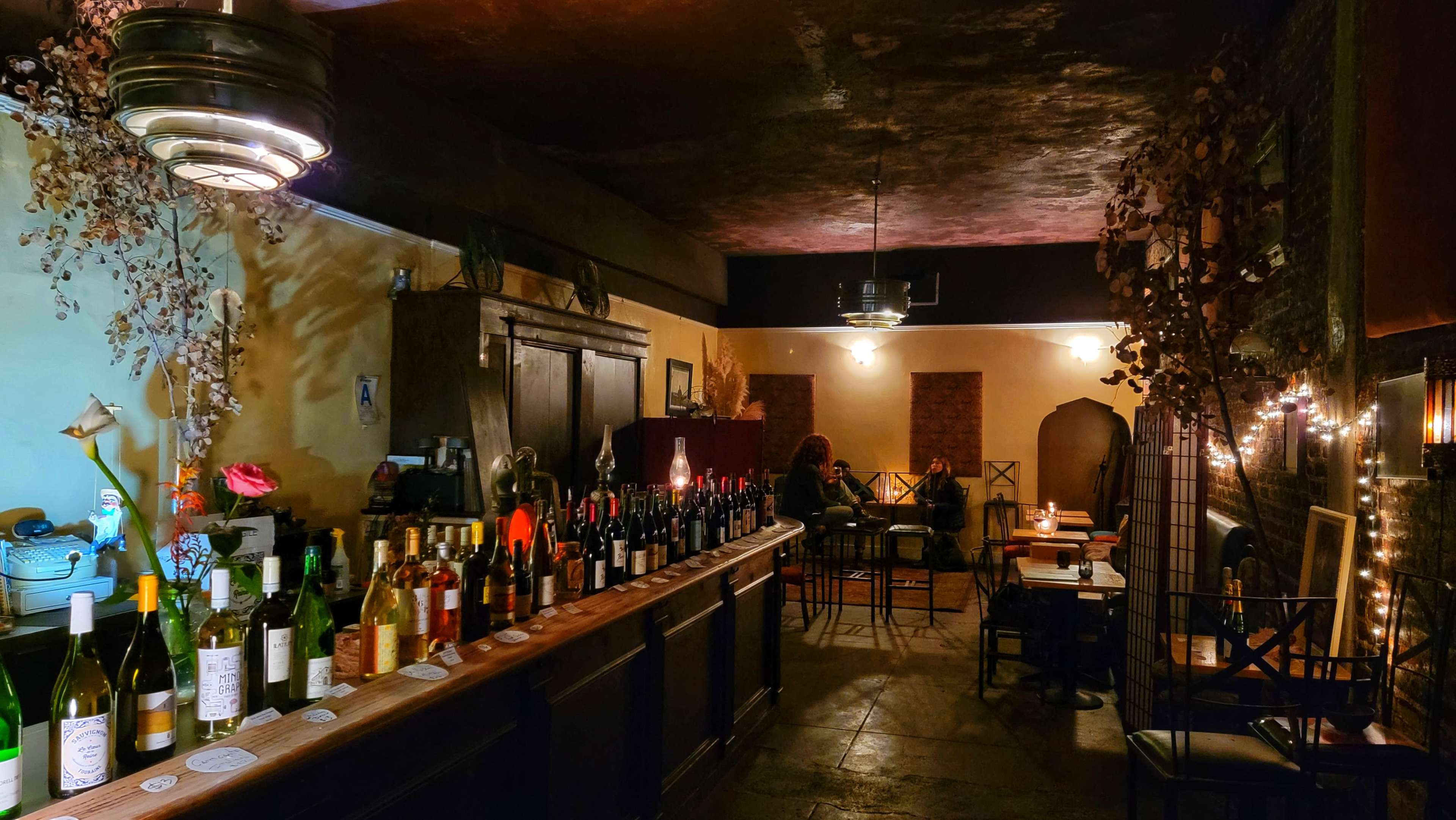 A dimly lit bar area features a long wooden counter with an array of bottles and tables in the background.