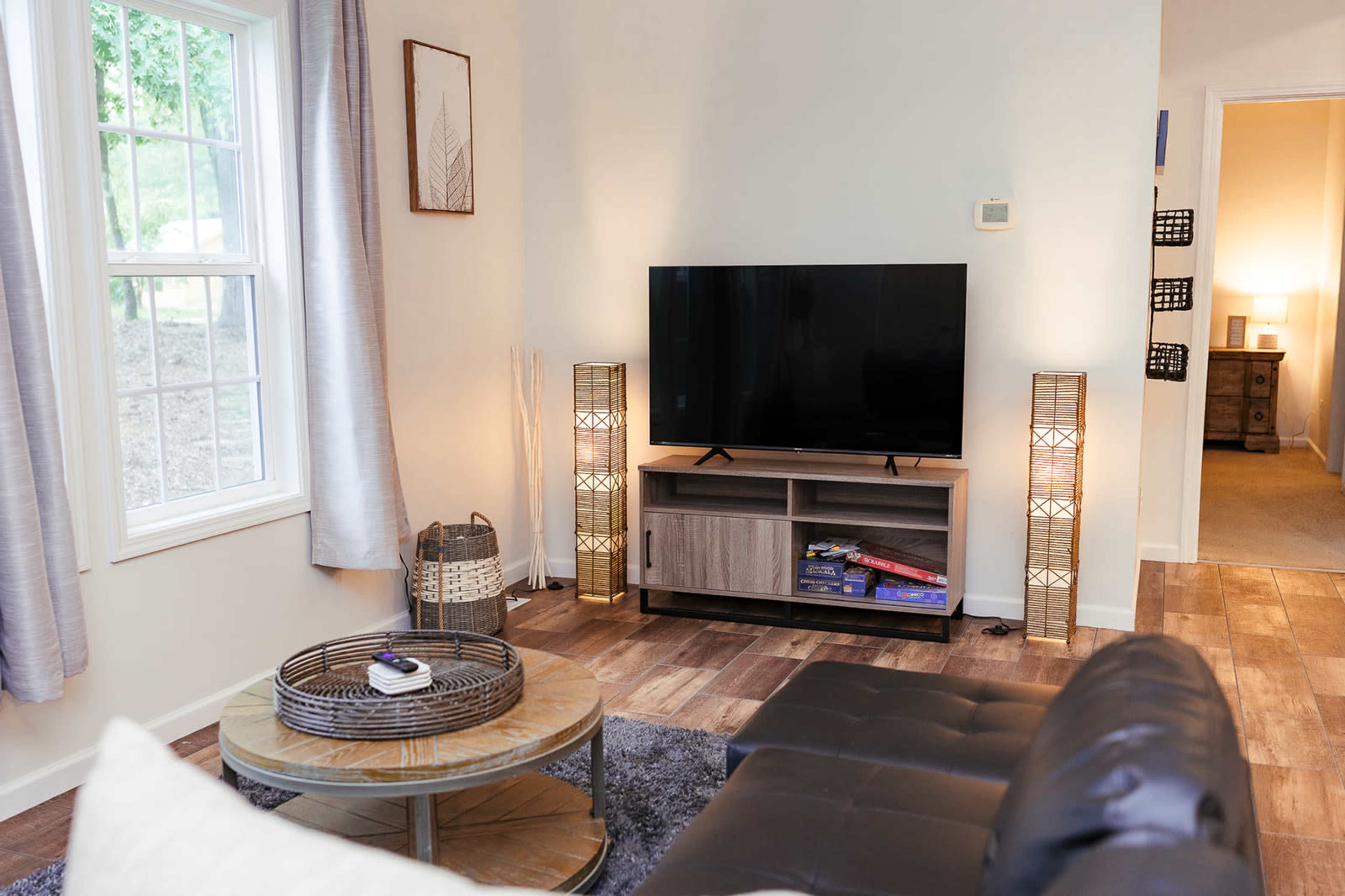 A living room features a television on a wooden stand, flanked by two decorative light fixtures, with a coffee table in front and a gray rug underfoot.