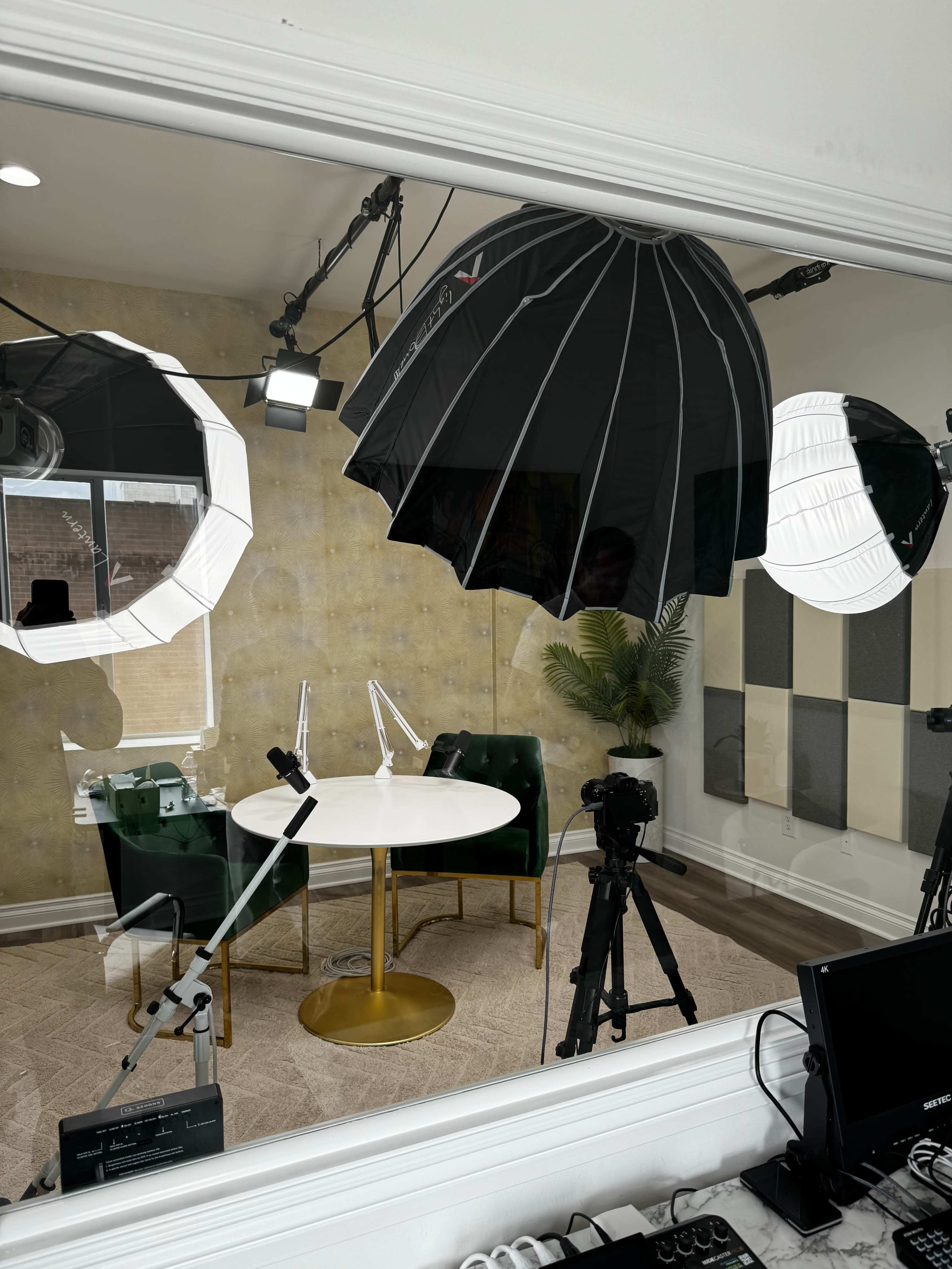 A recording studio setup with two green chairs, a round table, lighting equipment, and cameras arranged around the space.