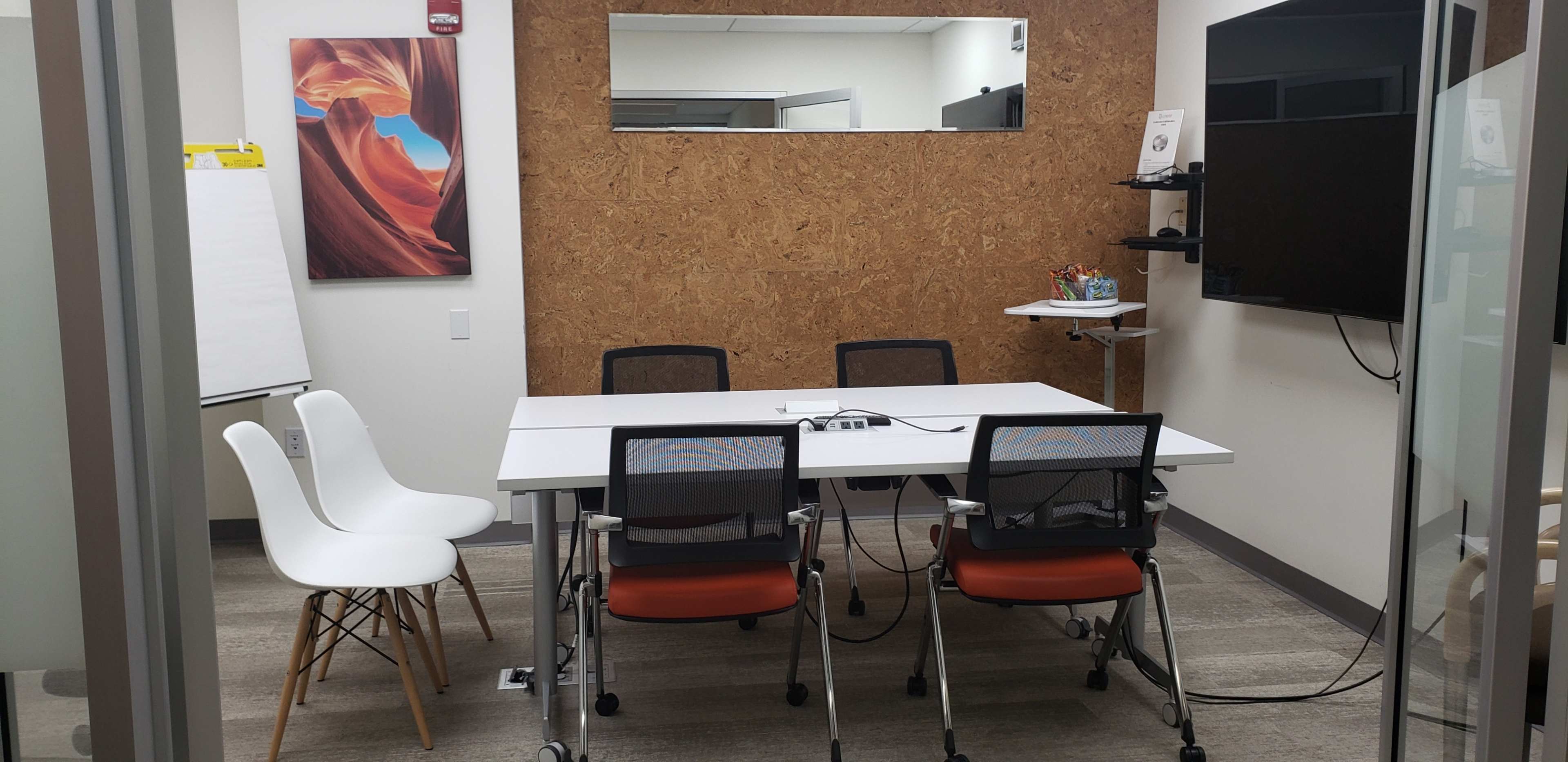 A conference room features a white table surrounded by four chairs, a large wall-mounted screen, and a corkboard backdrop.