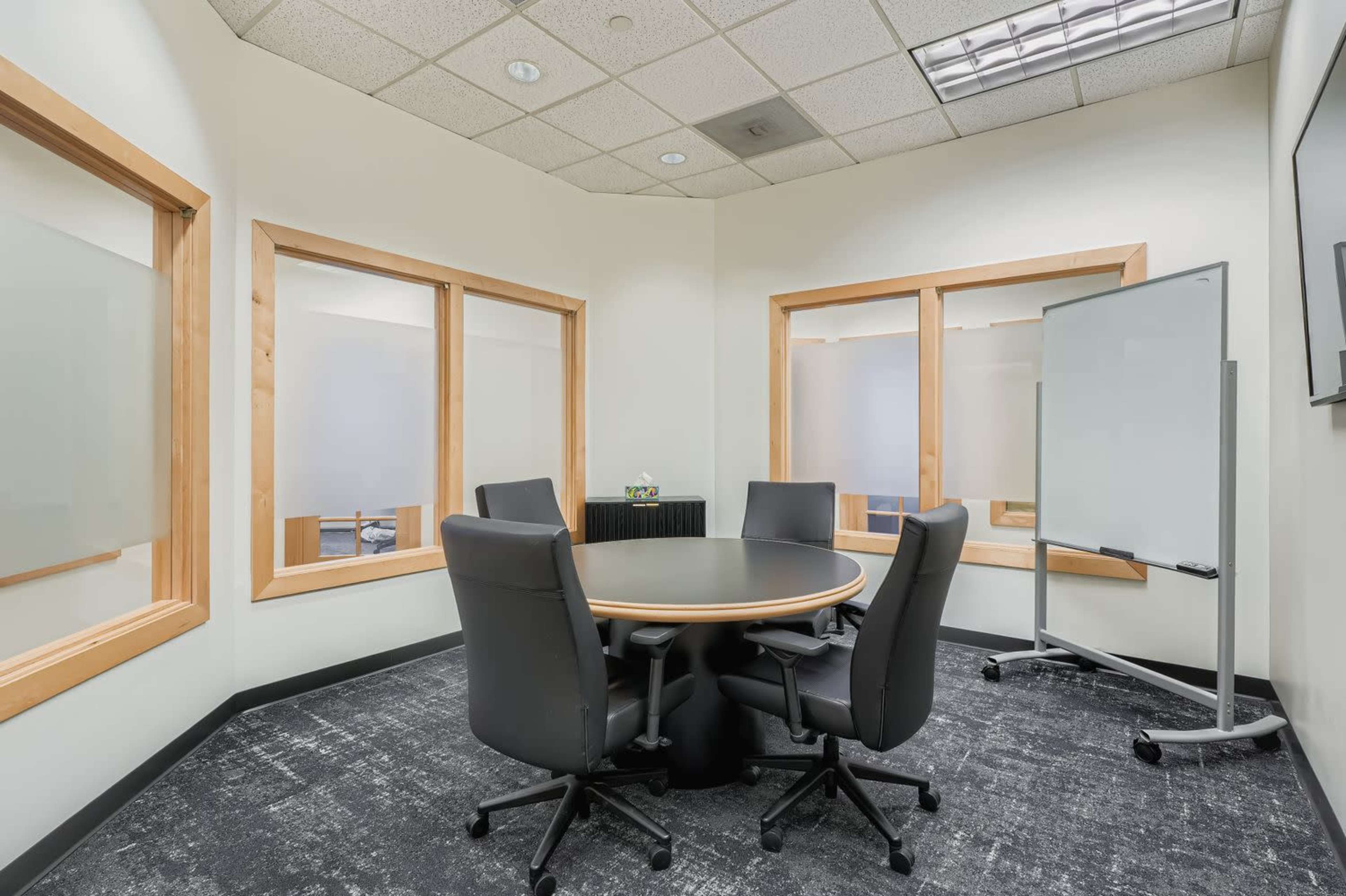 The image shows a conference room with a round table and four black chairs, surrounded by glass windows and a whiteboard.