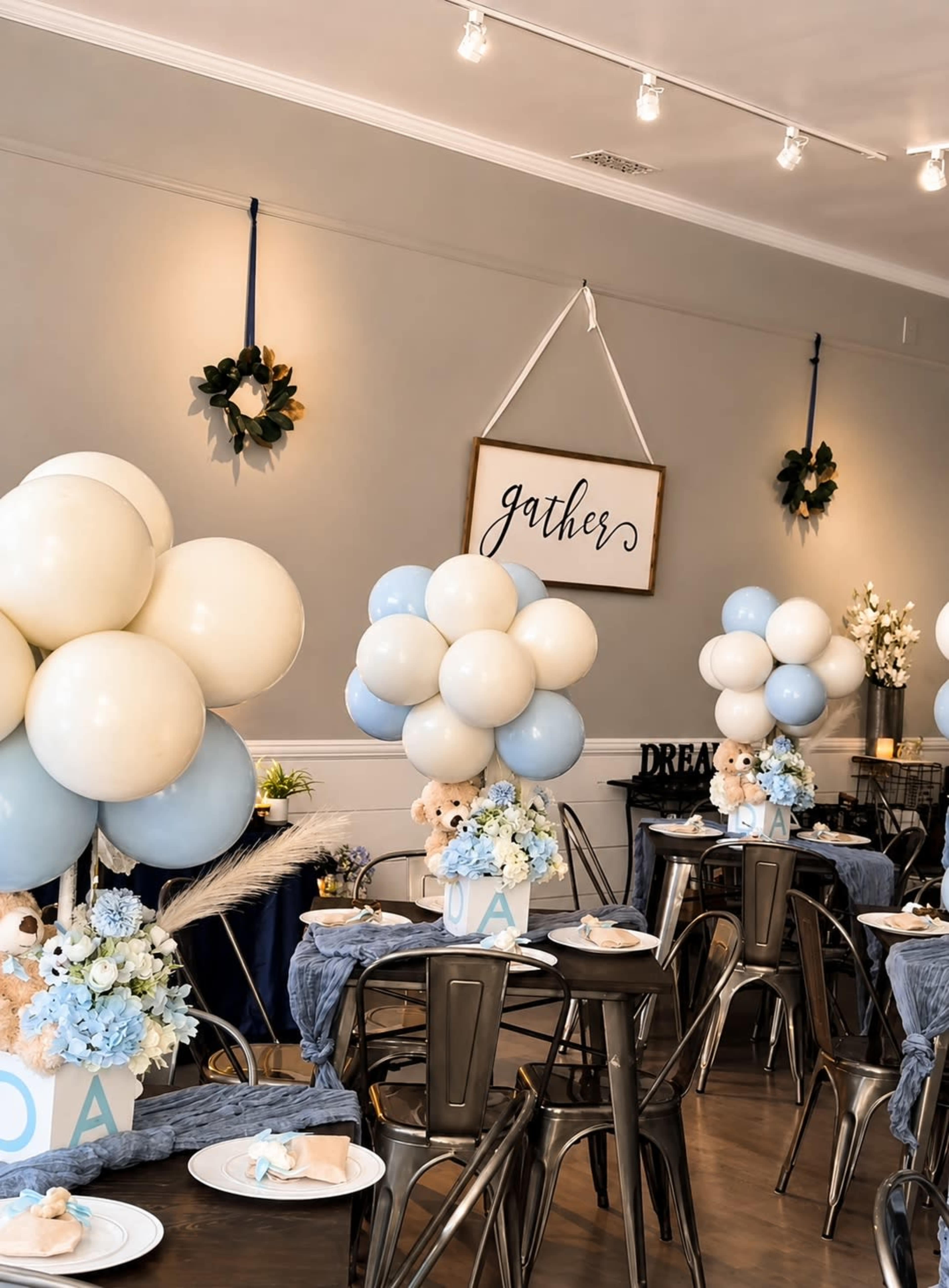 The image shows a beautifully decorated event space with tables set for a celebration, featuring arrangements of blue and white balloons, floral centerpieces, and a sign that reads "gathers."
