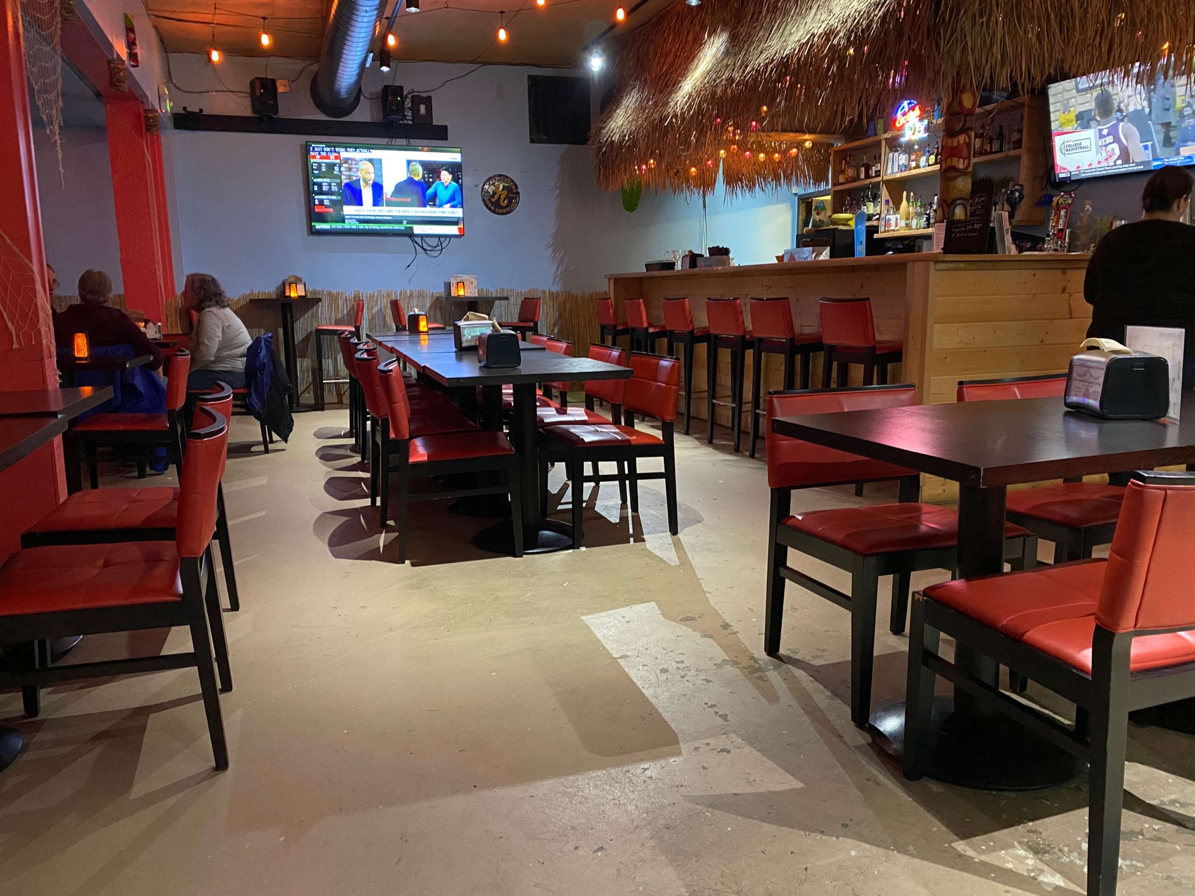 An empty restaurant interior featuring black tables and red chairs, with a bar area and multiple televisions displaying content.