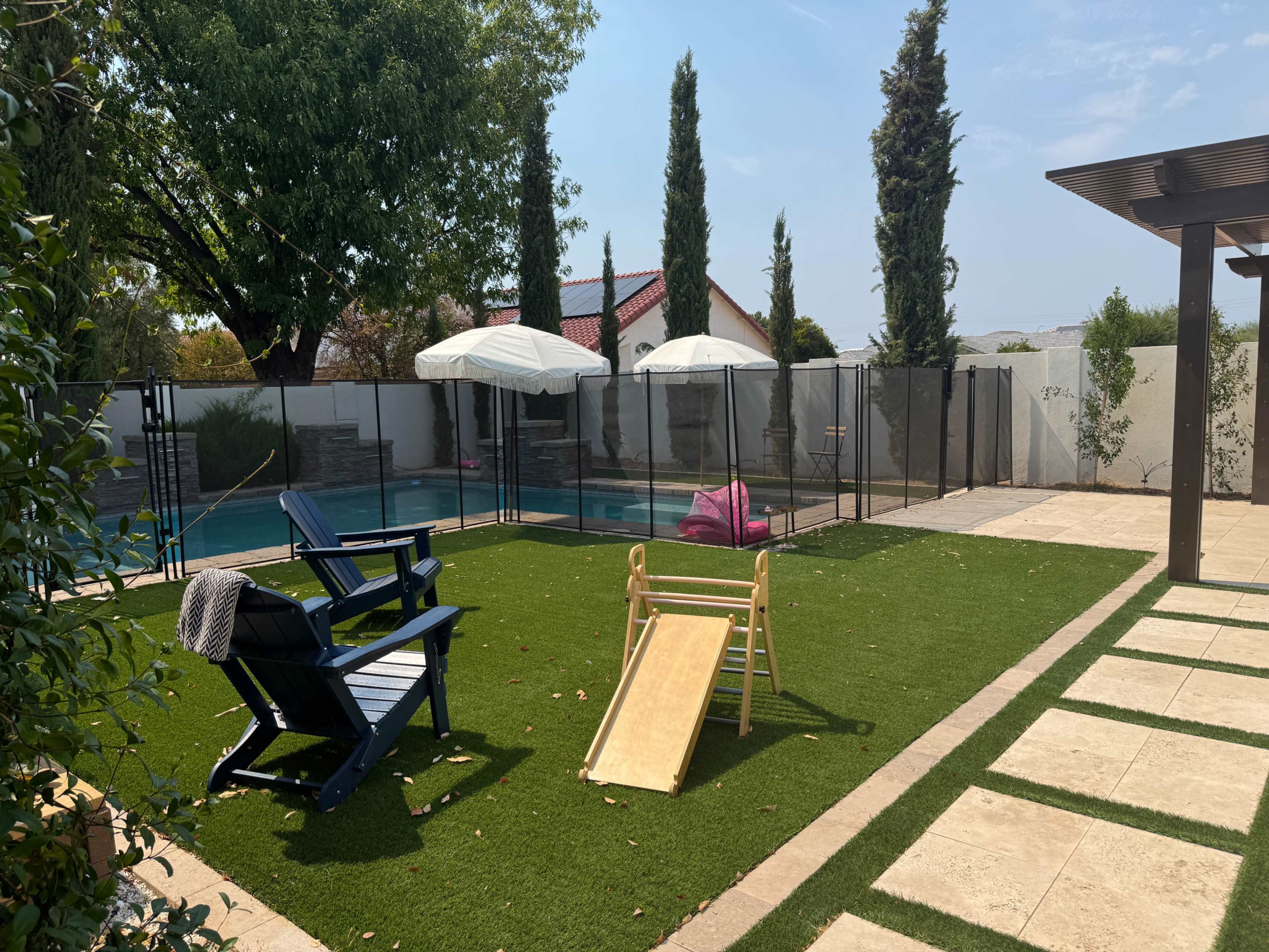 The image shows a backyard with a swimming pool, lounge chairs, a child’s slide, and a safety fence around the pool area, all surrounded by green grass.