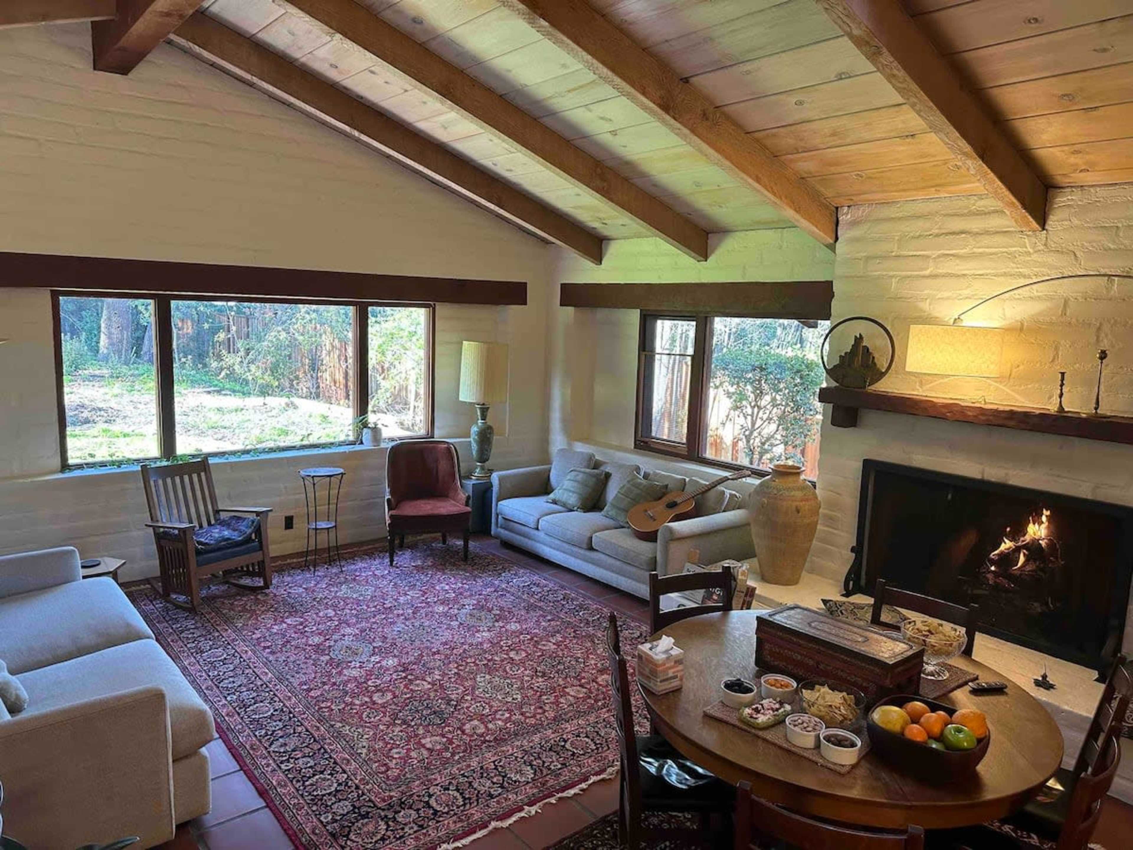 A cozy living room with a fireplace, a large area rug, a round wooden dining table, and windows overlooking a natural landscape.