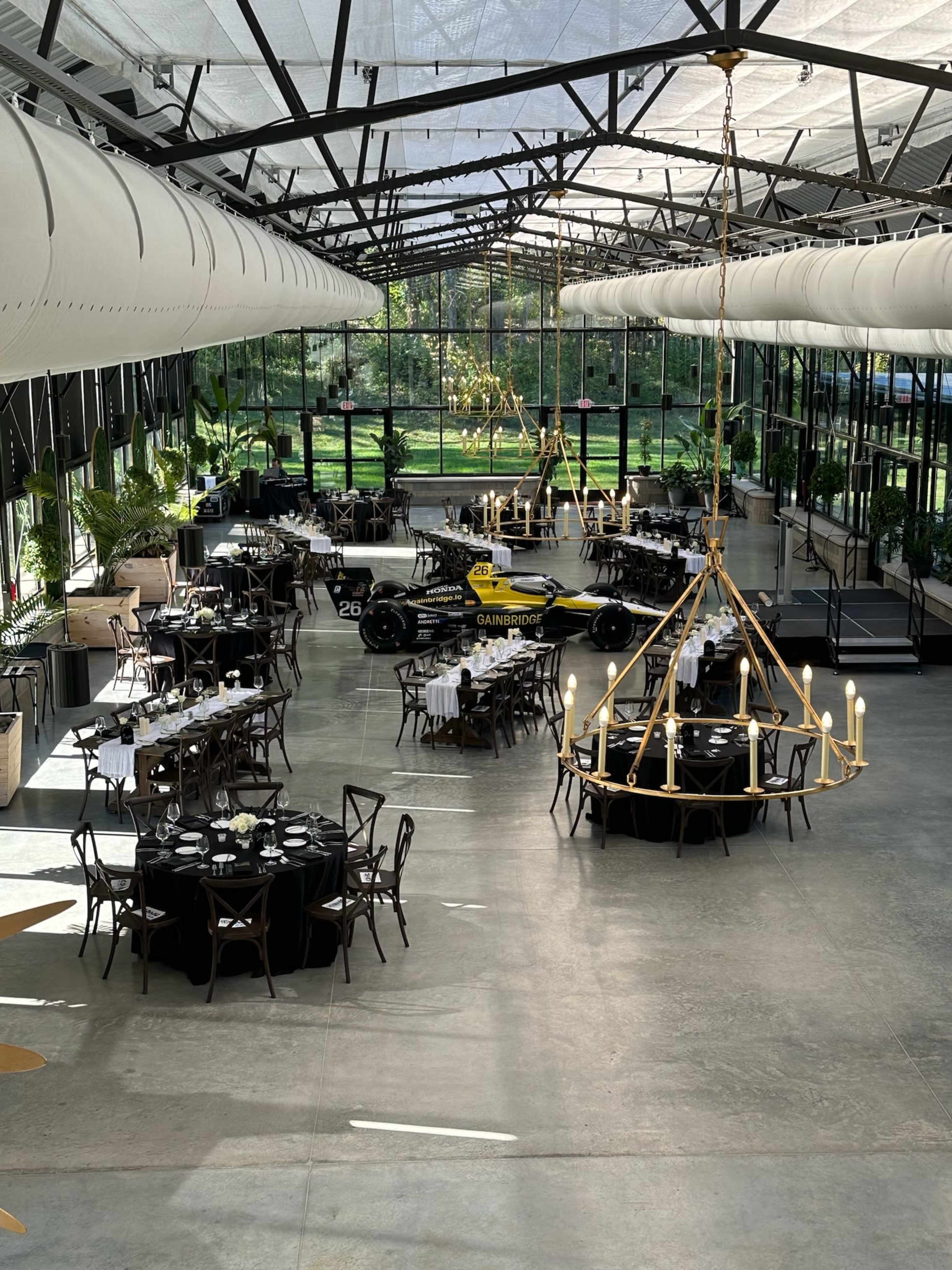 The image shows a spacious, modern event space featuring dining tables, a race car on display, and large windows that allow natural light to fill the room.