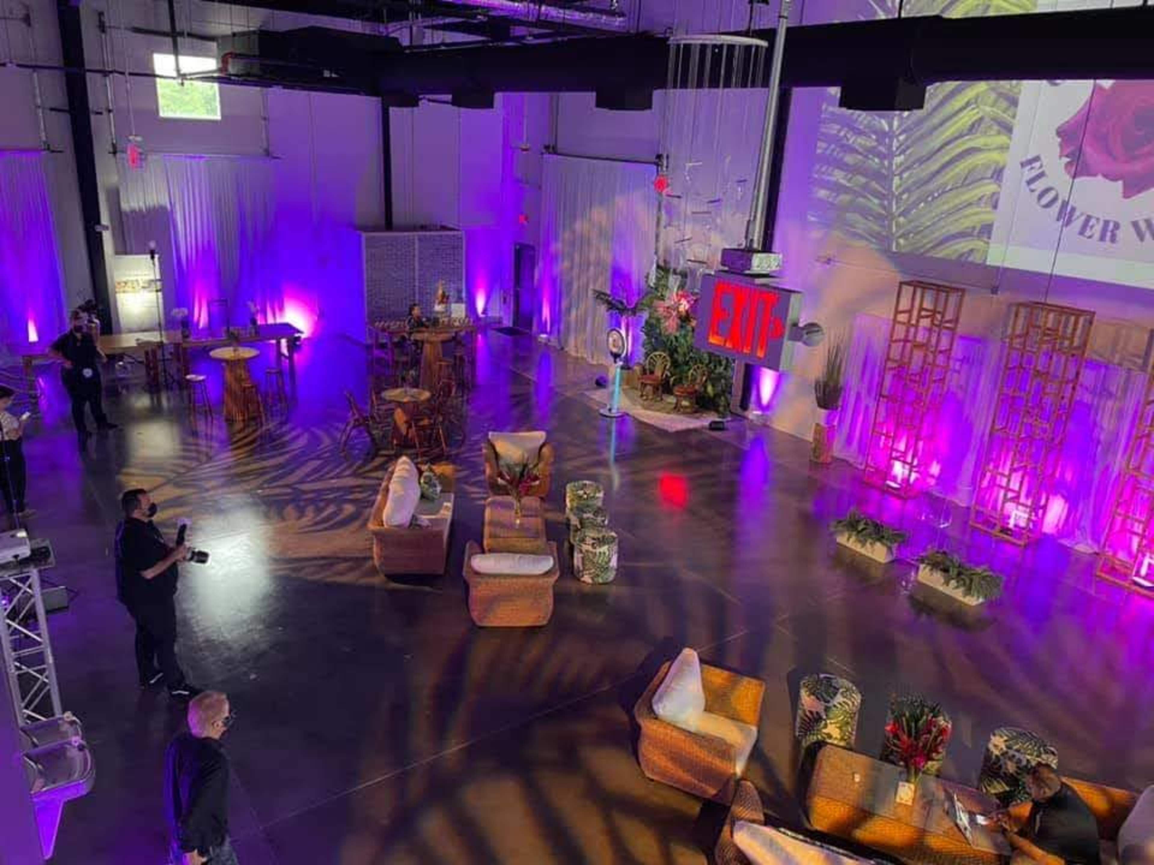 The image shows an event space with a modern setup featuring wicker furniture, decorative plants, and colorful lighting, including purple hues and an illuminated "EXIT" sign.