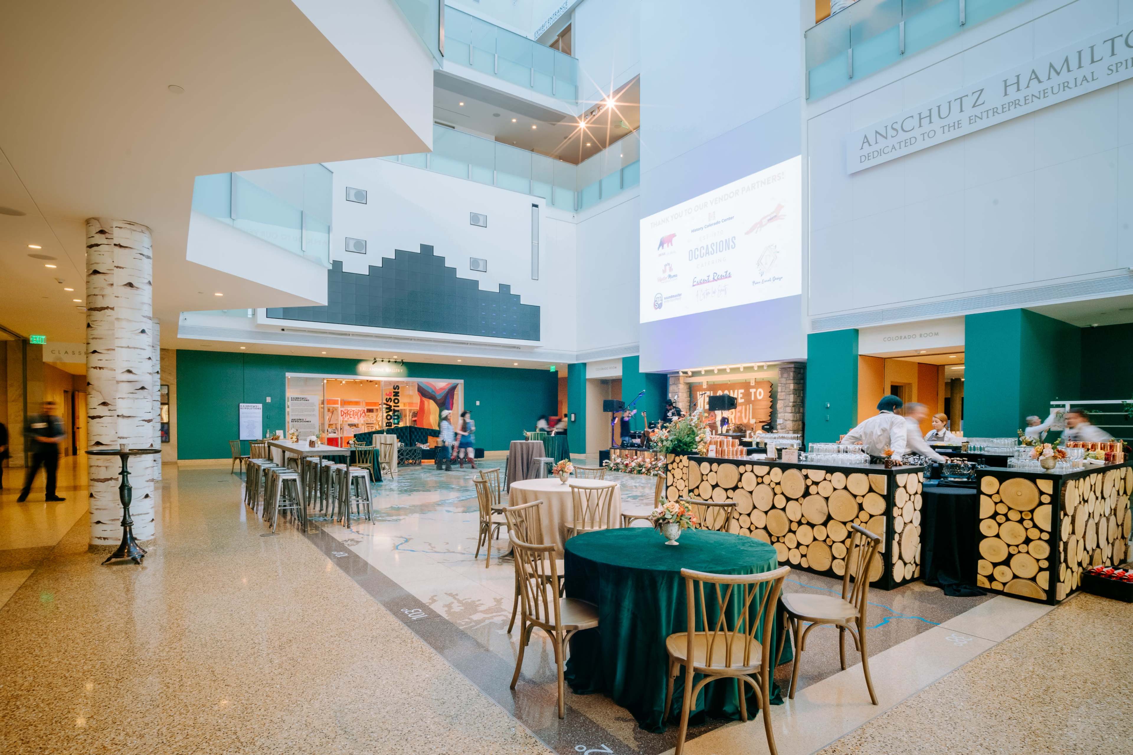 A spacious indoor venue features tables set with green tablecloths and a buffet area, all surrounded by modern architectural design and decorative accents.