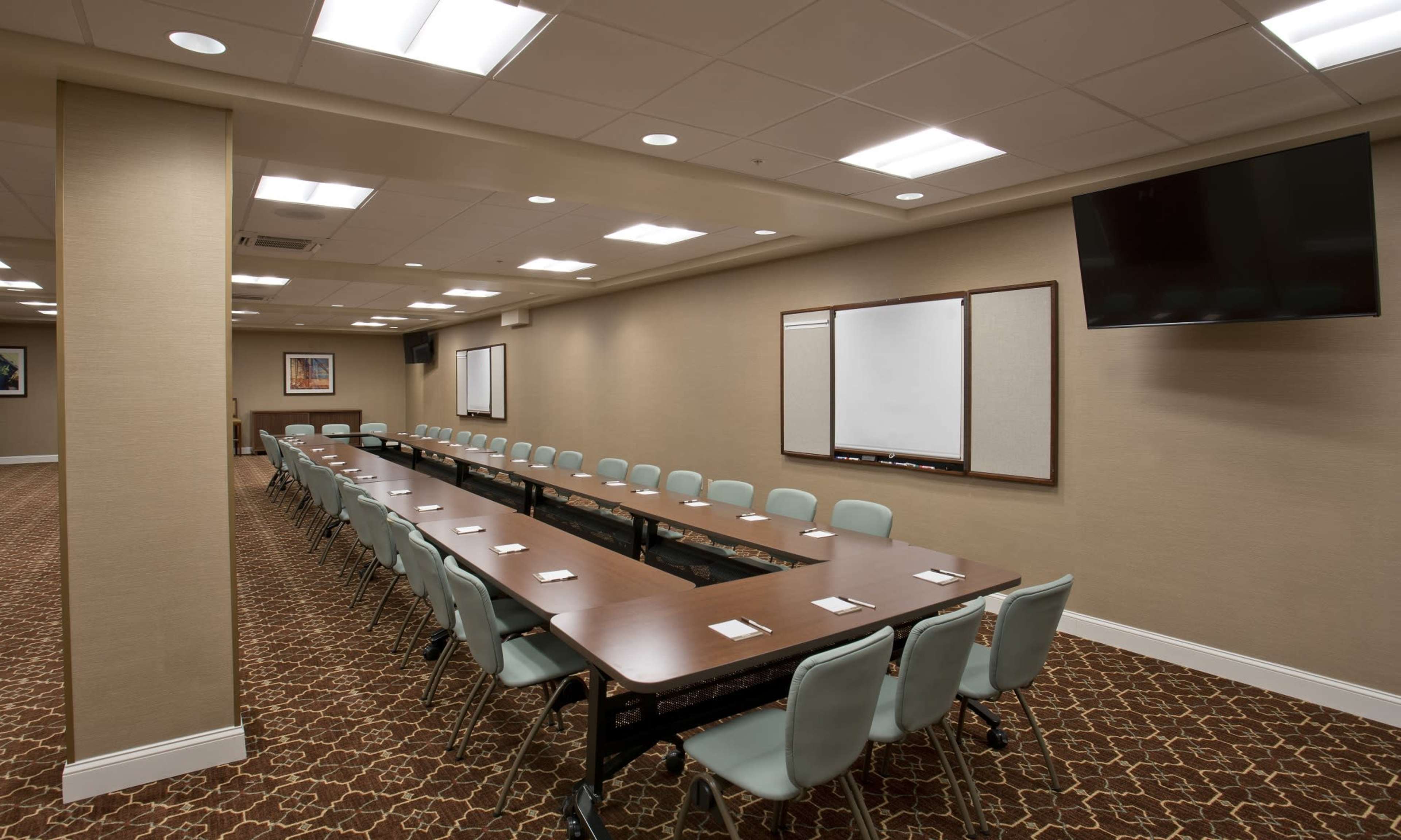A large meeting room features a long table with multiple chairs arranged around it, complemented by a whiteboard and a television mounted on the wall.