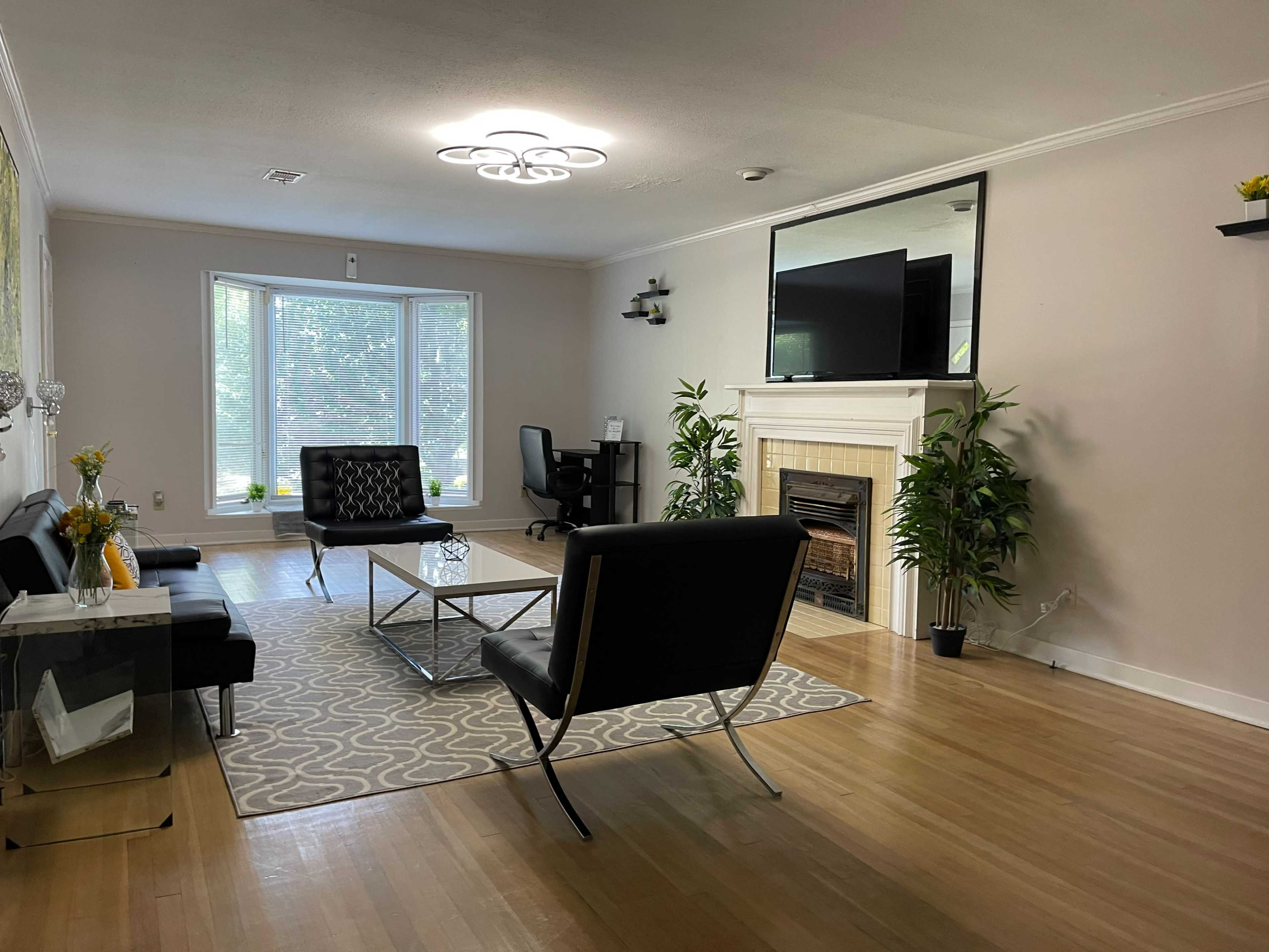 A spacious living room featuring a fireplace, two black chairs, a coffee table, and a work desk near a large window.