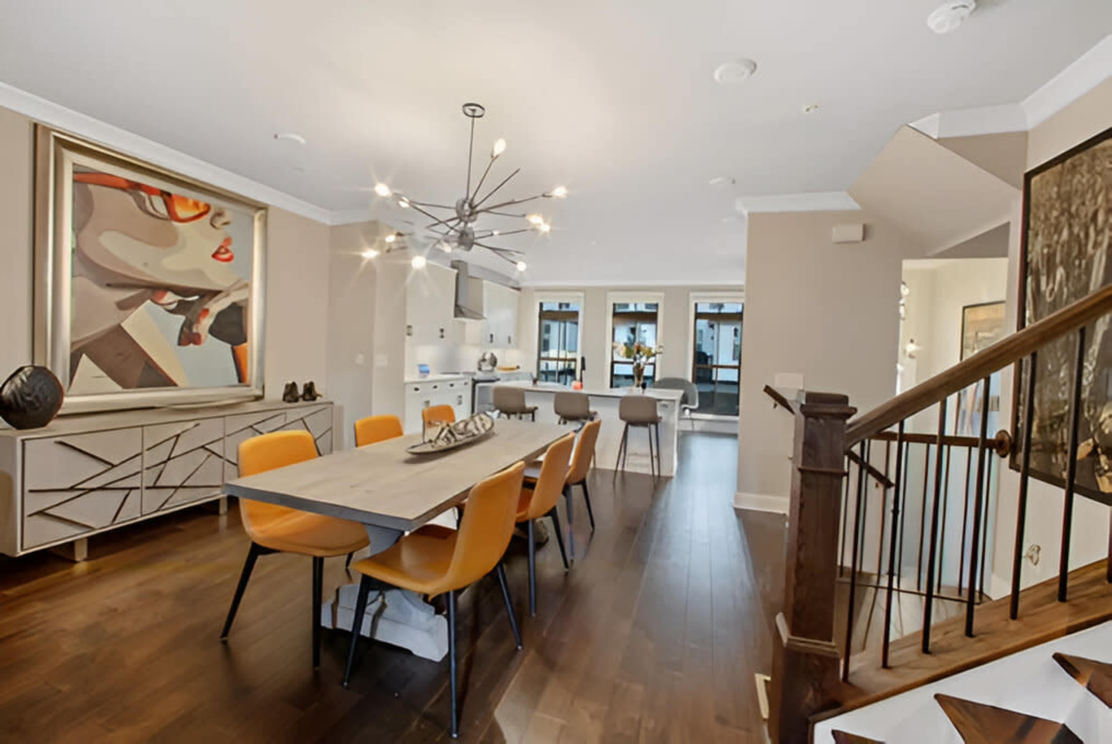 A modern dining area with a long table surrounded by orange chairs, a large painting on the wall, and a staircase in the corner leading to another level.