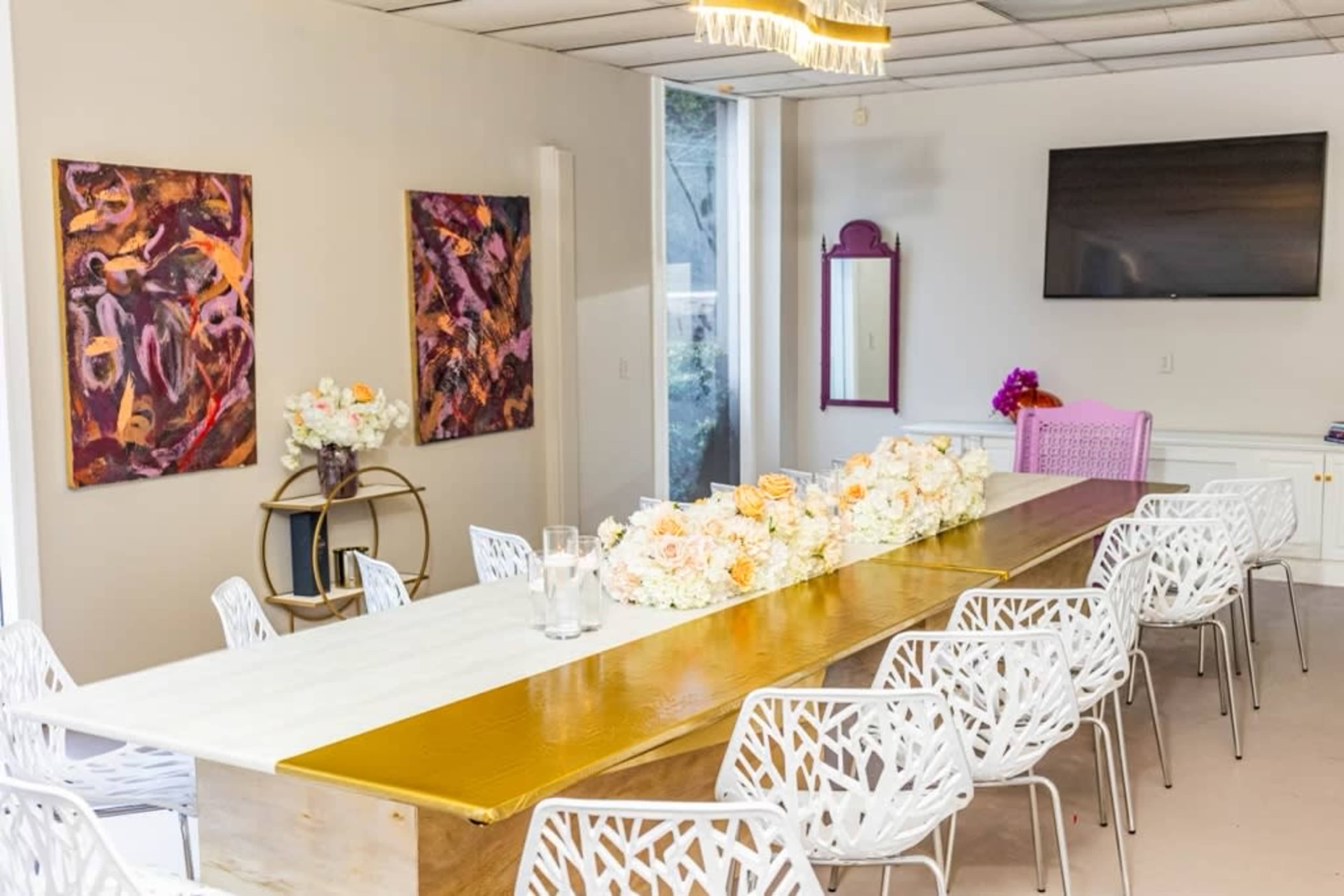 A long wooden table is set in a well-lit room with white chairs, floral centerpieces, and two colorful paintings on the walls.