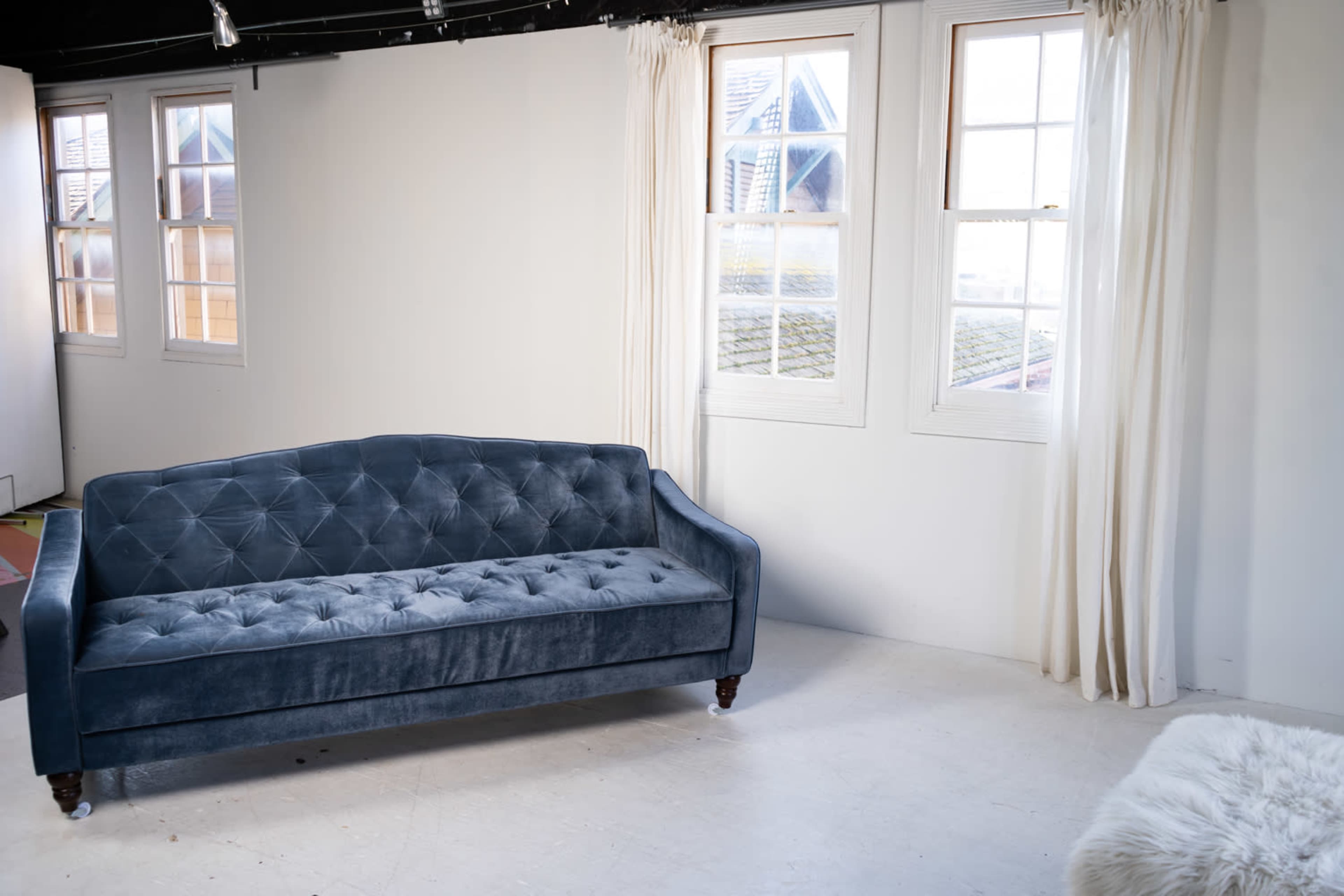 The image shows a blue velvet sofa with a tufted design placed against a white wall, illuminated by natural light coming through two windows with white curtains.