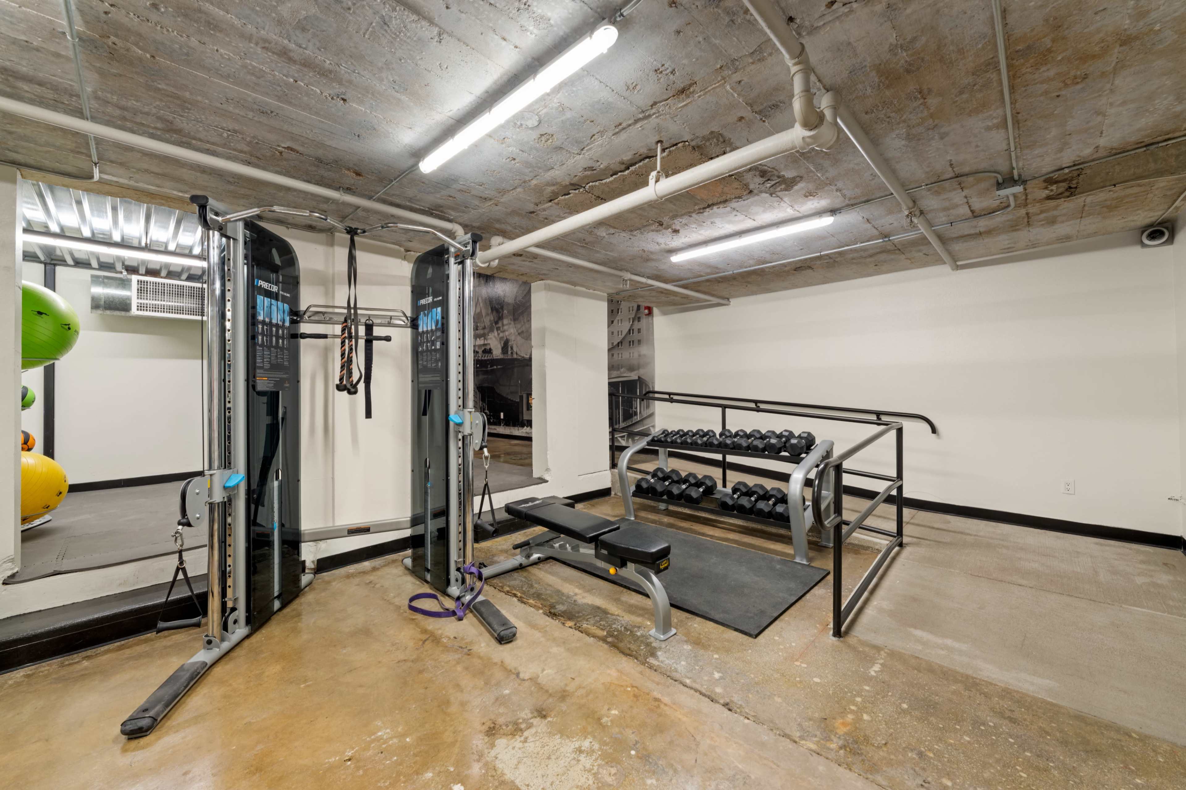 A gym area featuring a cable machine, a weight bench, a rack of dumbbells, and a concrete floor with overhead lighting.