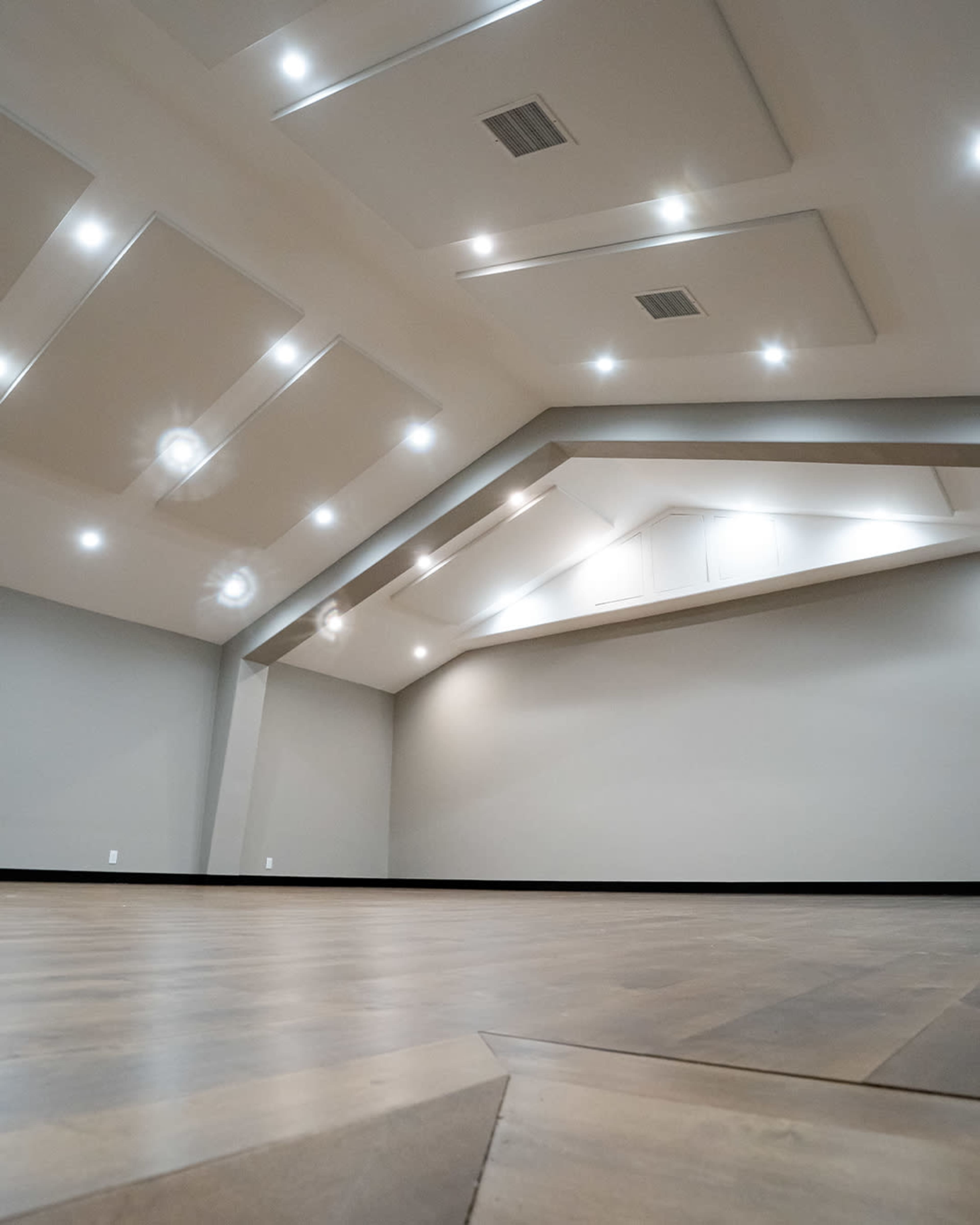 The image shows an empty room with a high ceiling, wooden flooring, and multiple recessed lights.