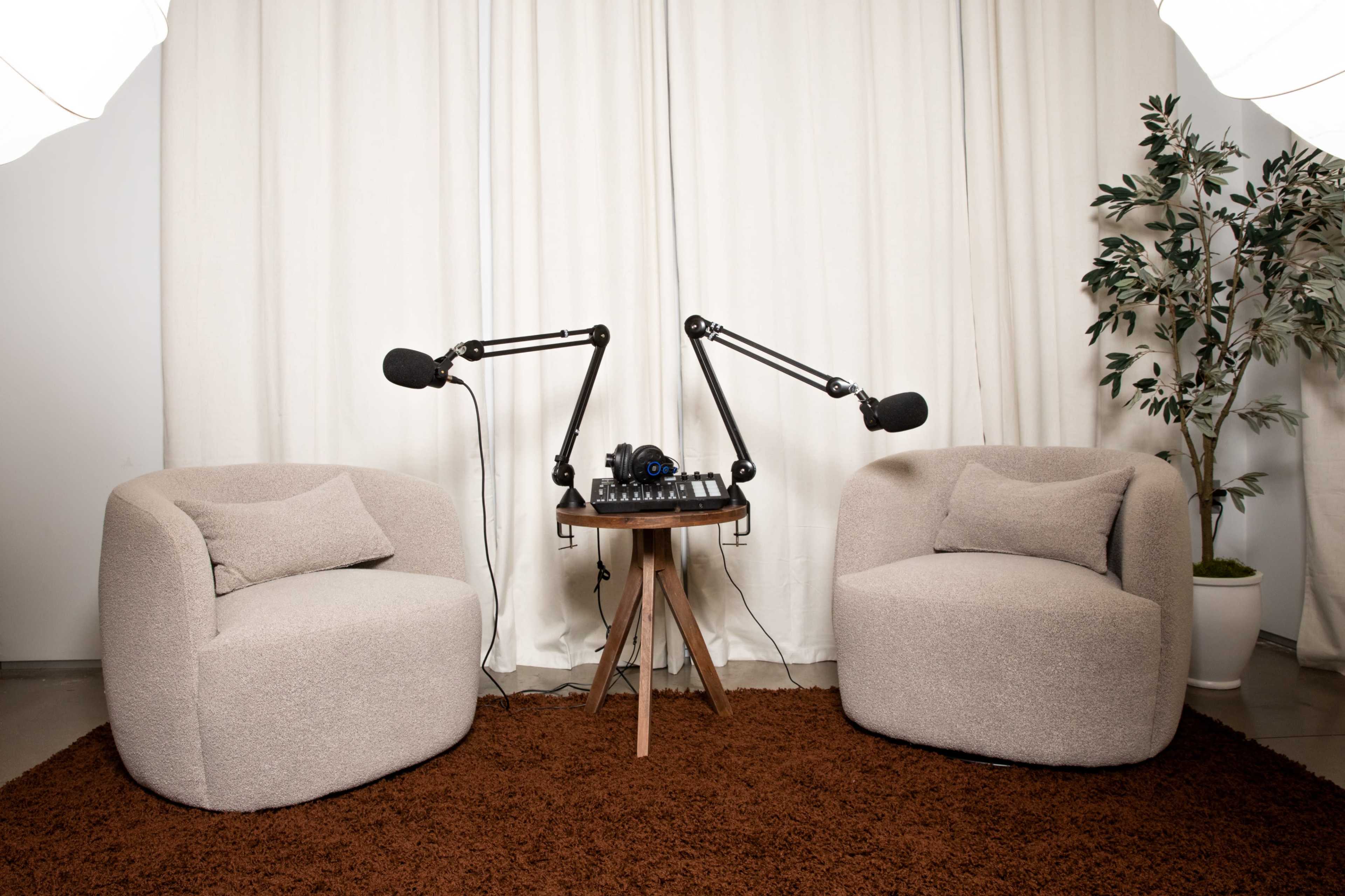 The image shows two upholstered chairs facing a small table equipped with microphones and audio equipment, set against a backdrop of a white curtain and a plant.