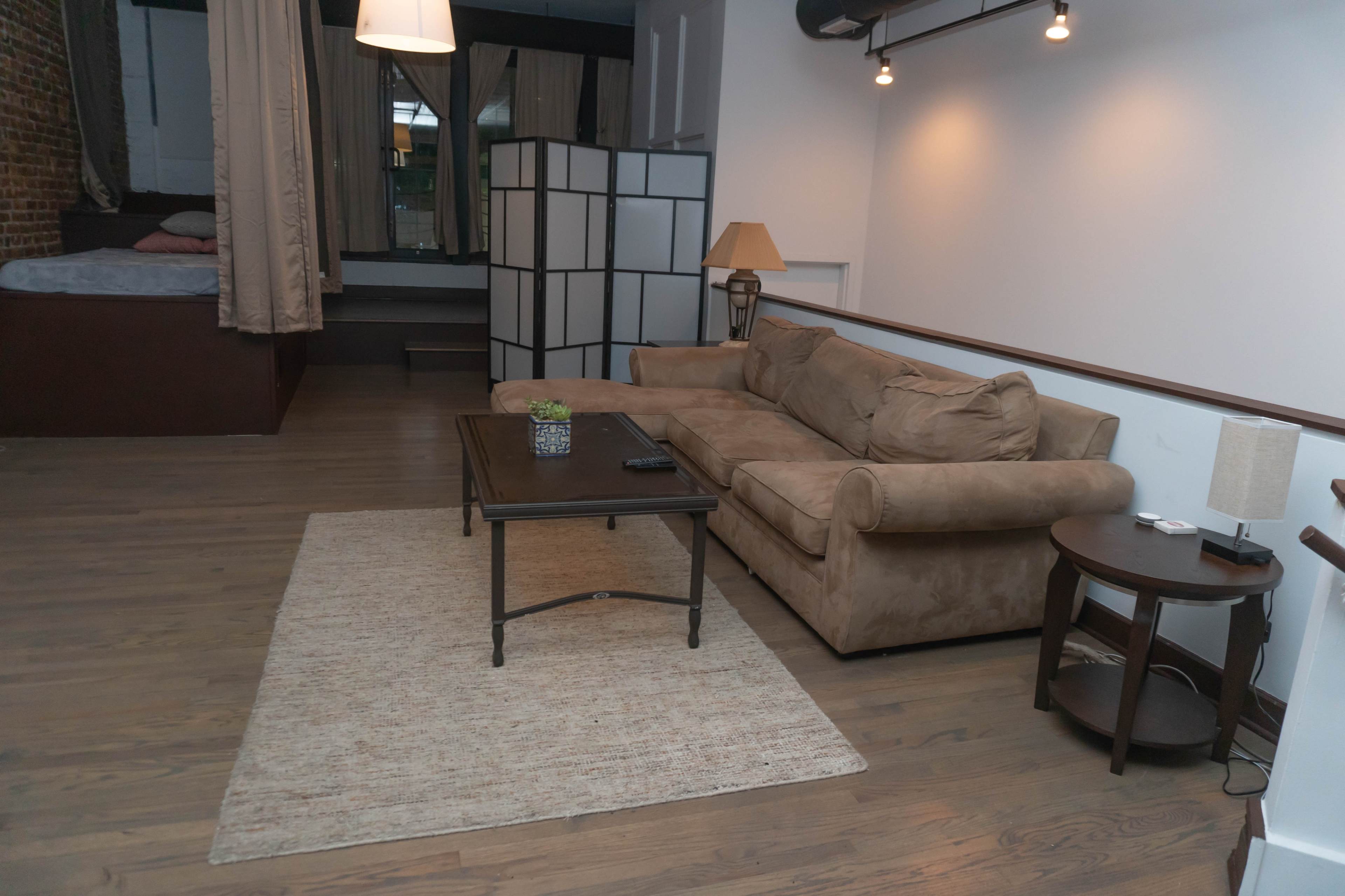 A living area furnished with a brown couch, a coffee table, a lamp, and a bed area separated by a screen.
