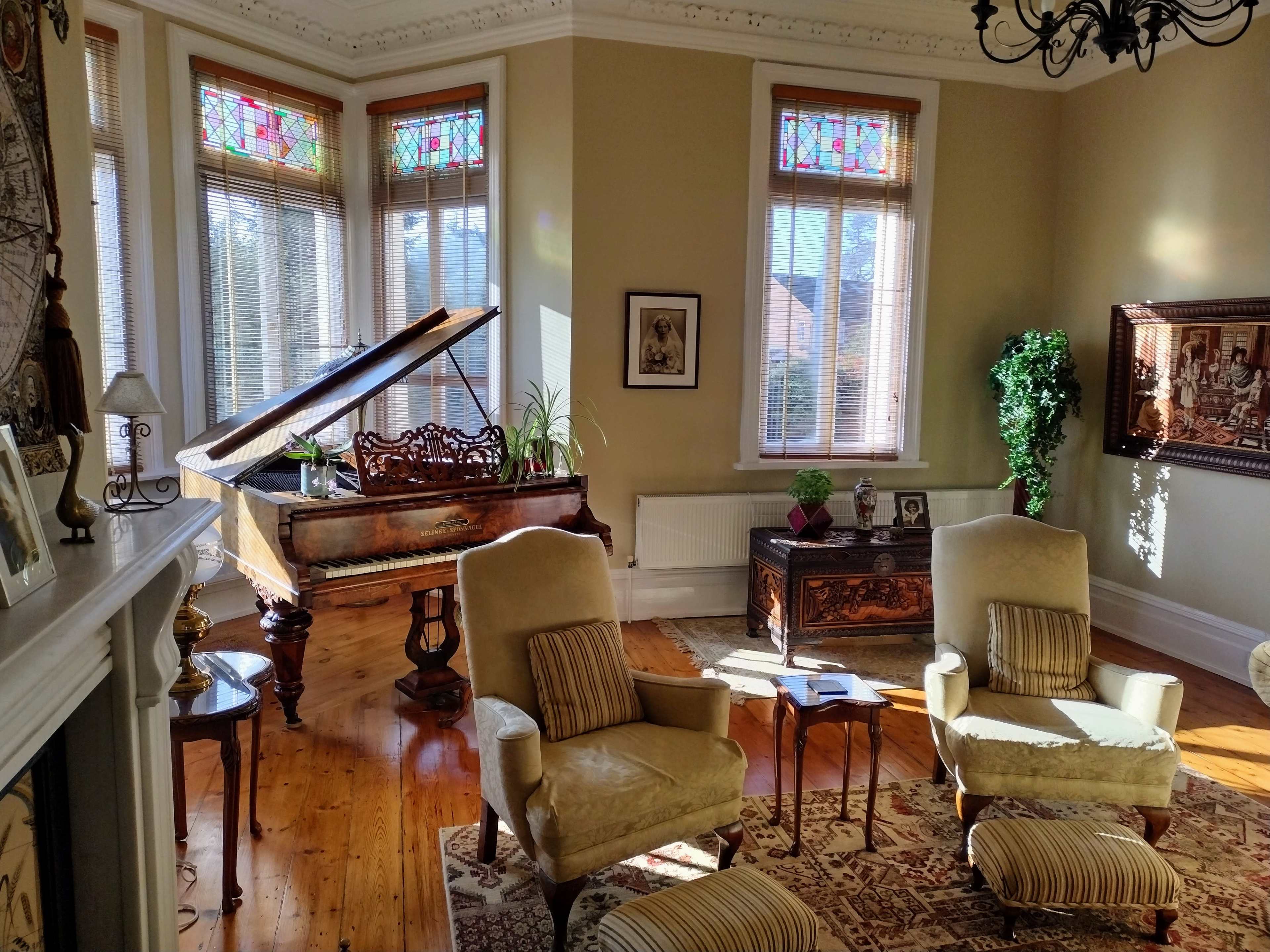 A sunlit living room featuring a piano, two upholstered chairs, a decorative rug, and stained glass windows.