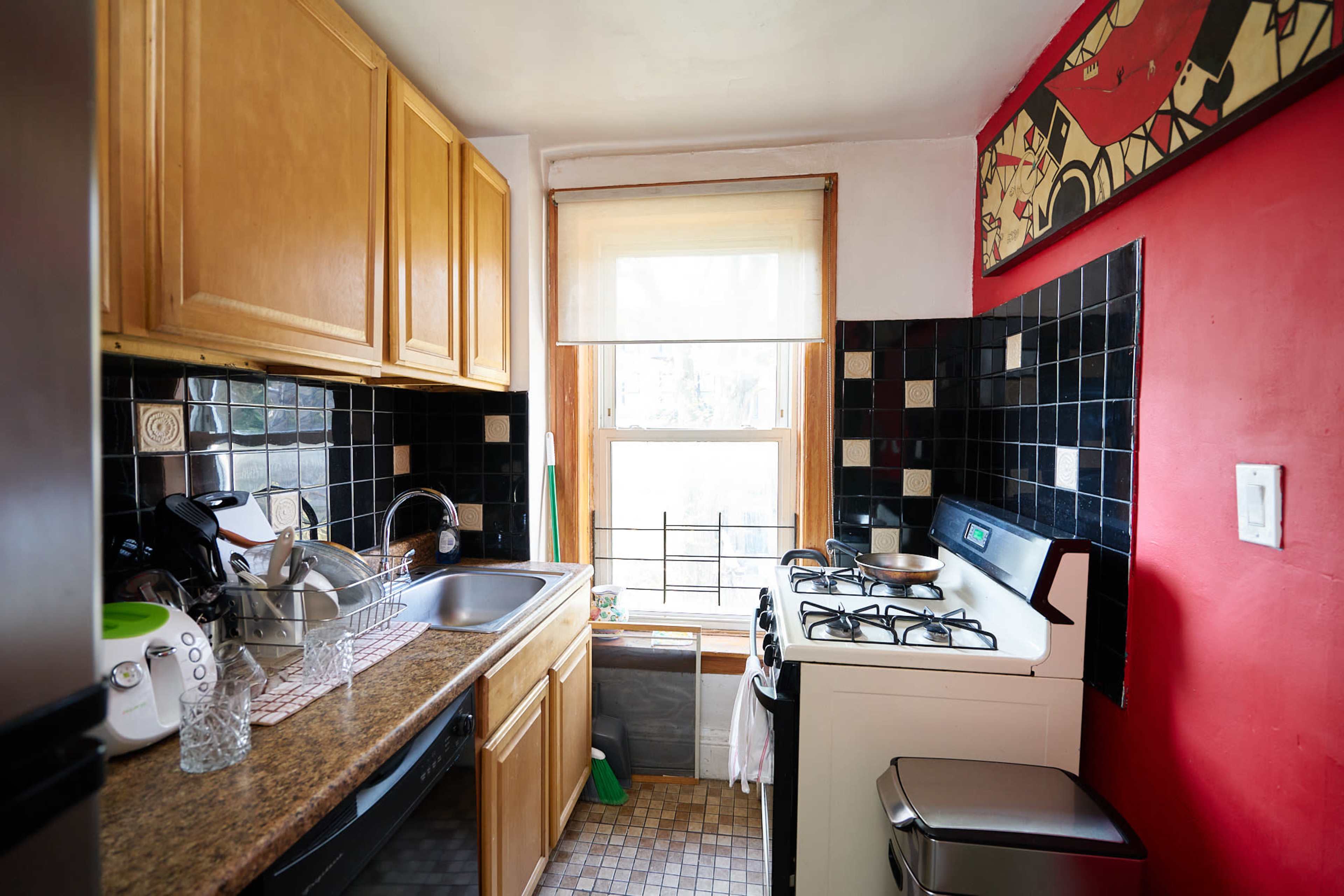 The image shows a compact kitchen with wooden cabinets, black and white tiled walls, and a stove next to a window.