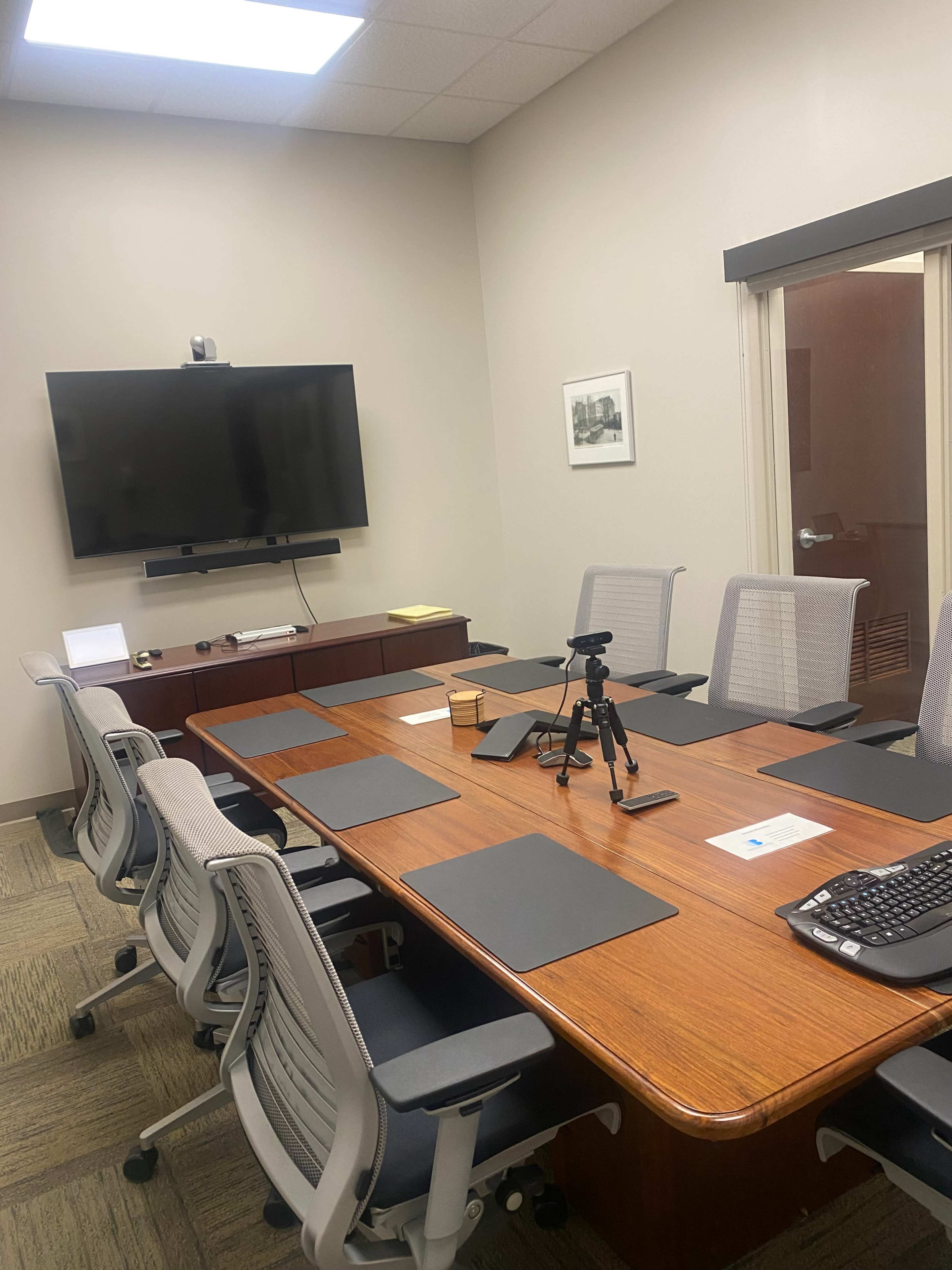 A conference room with a large wooden table surrounded by ergonomic chairs, equipped with a TV on the wall and a tripod on the table.