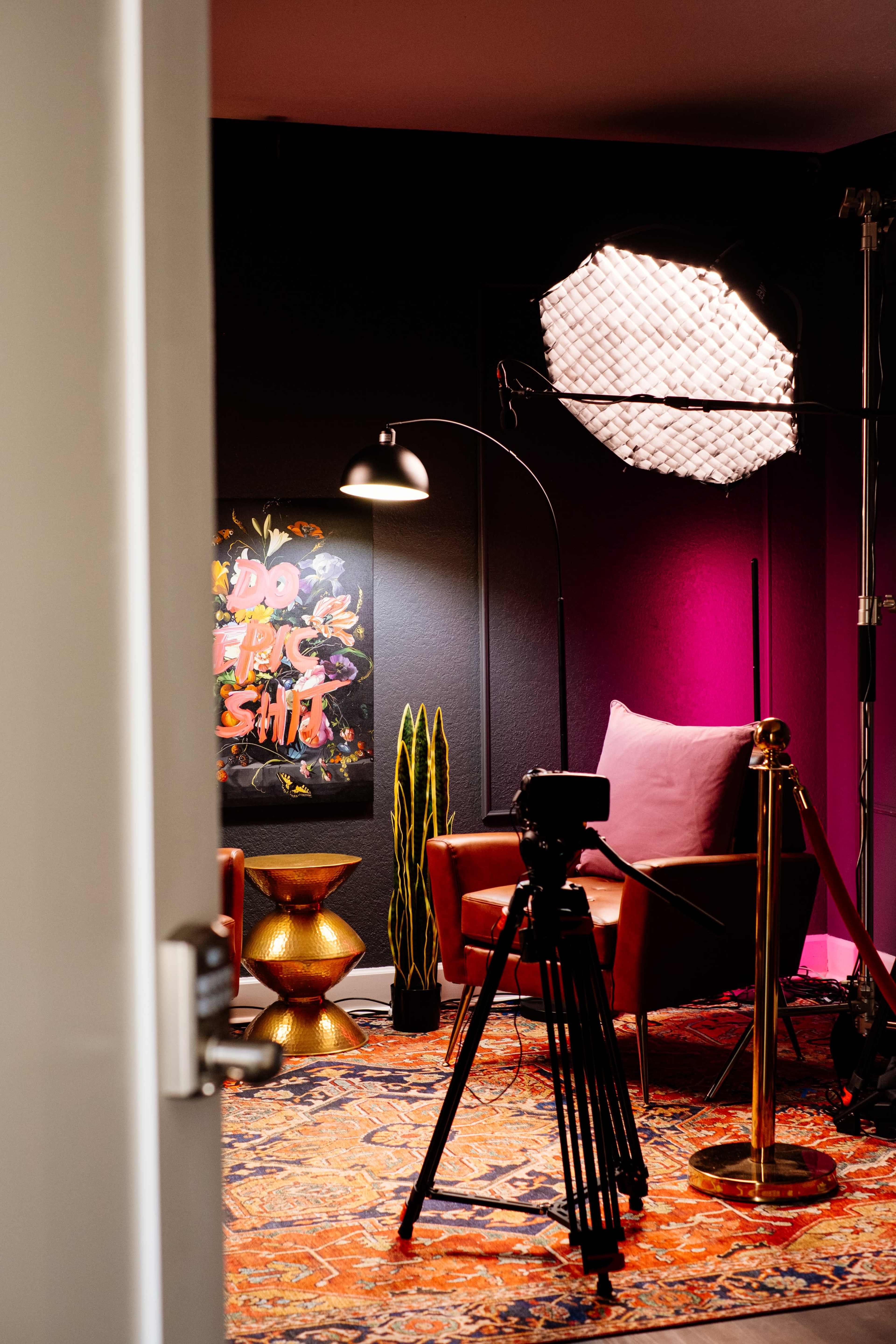 A well-lit recording studio features a camera on a tripod, a colorful rug, modern furniture, and vibrant artwork on the wall.