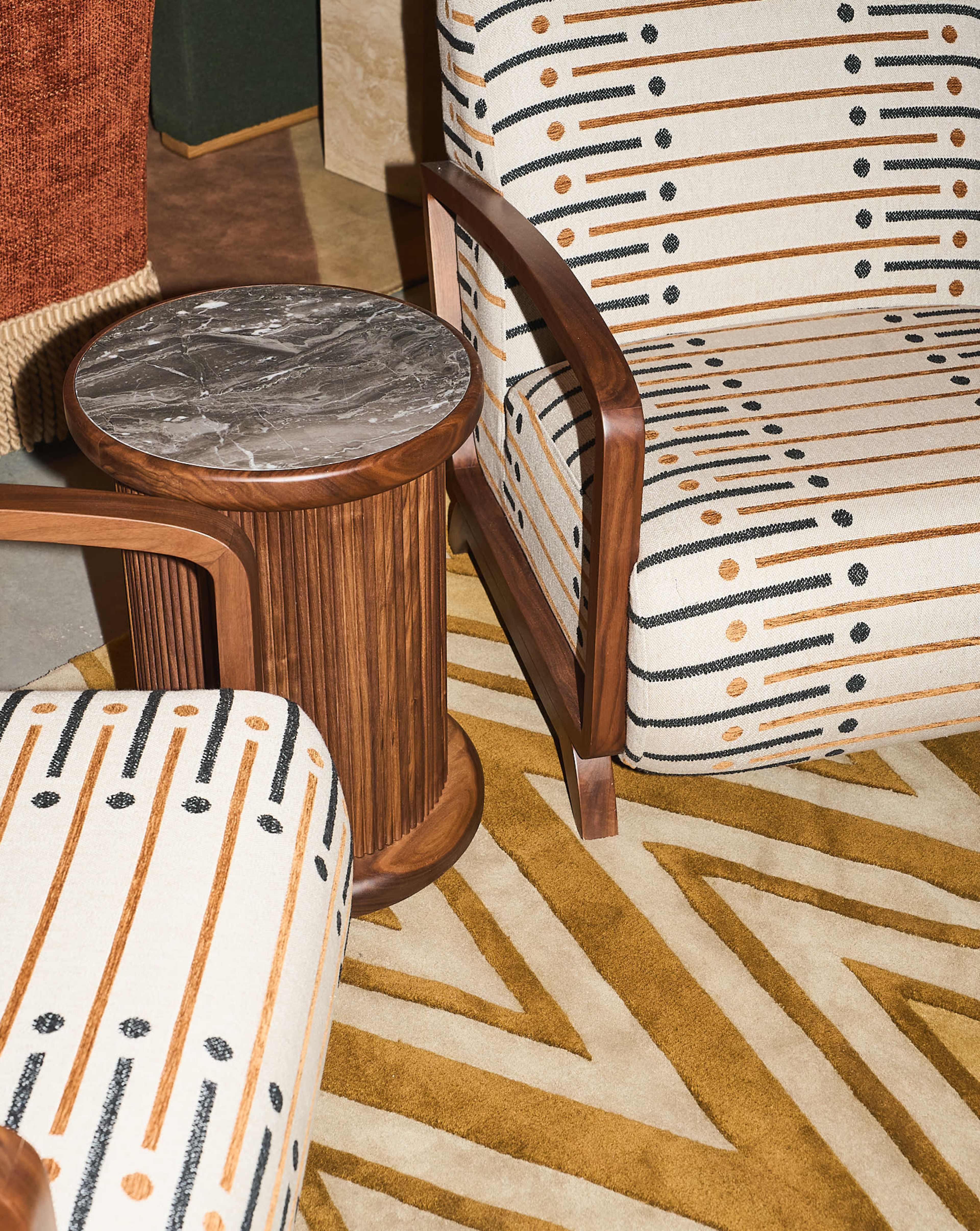 The image shows a stylish interior setup featuring two patterned chairs and a cylindrical wooden side table with a marble top, placed on a geometric area rug.