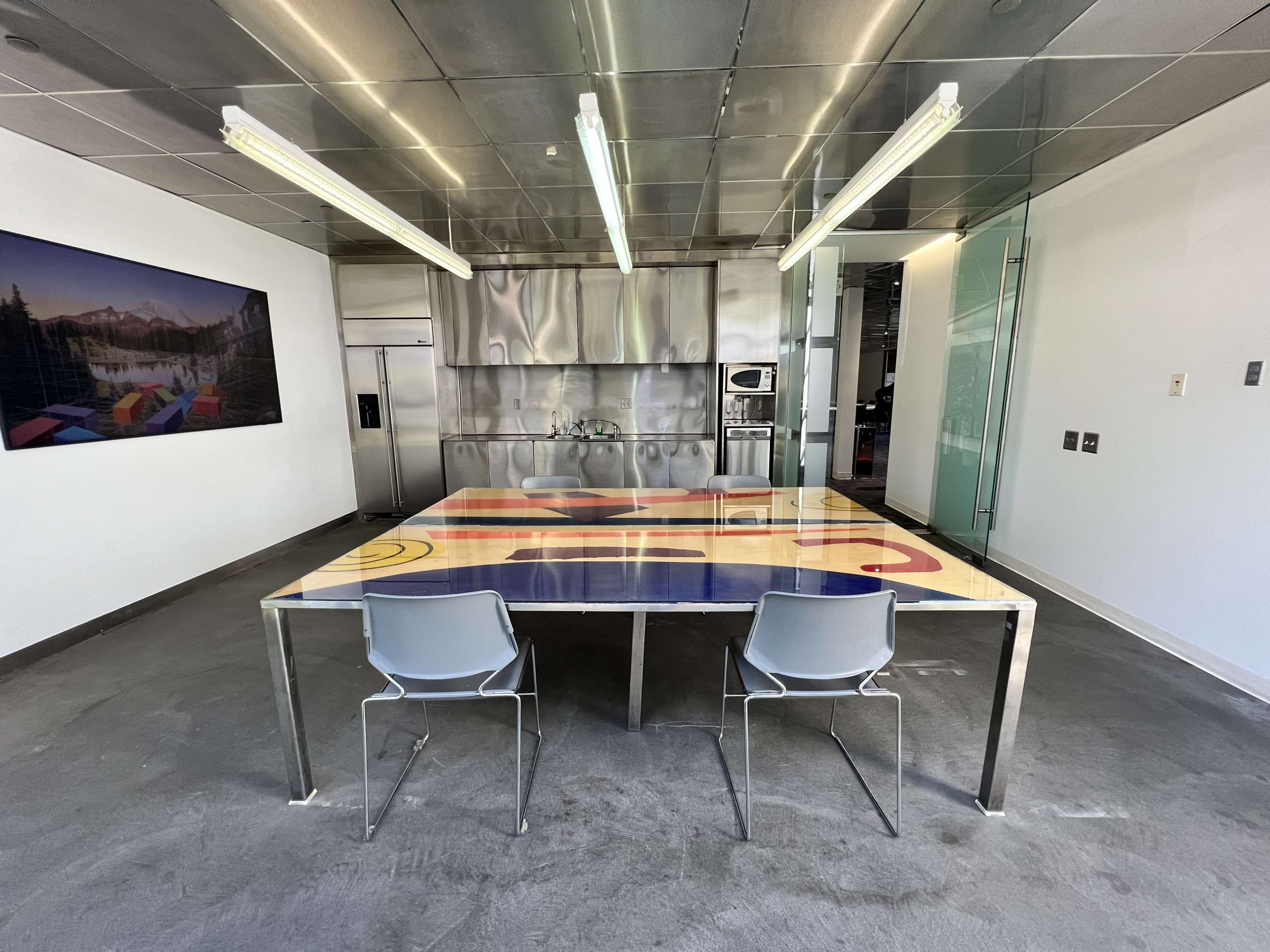 A modern kitchen features a stainless steel interior and a large table with a colorful design, accompanied by two metal chairs.