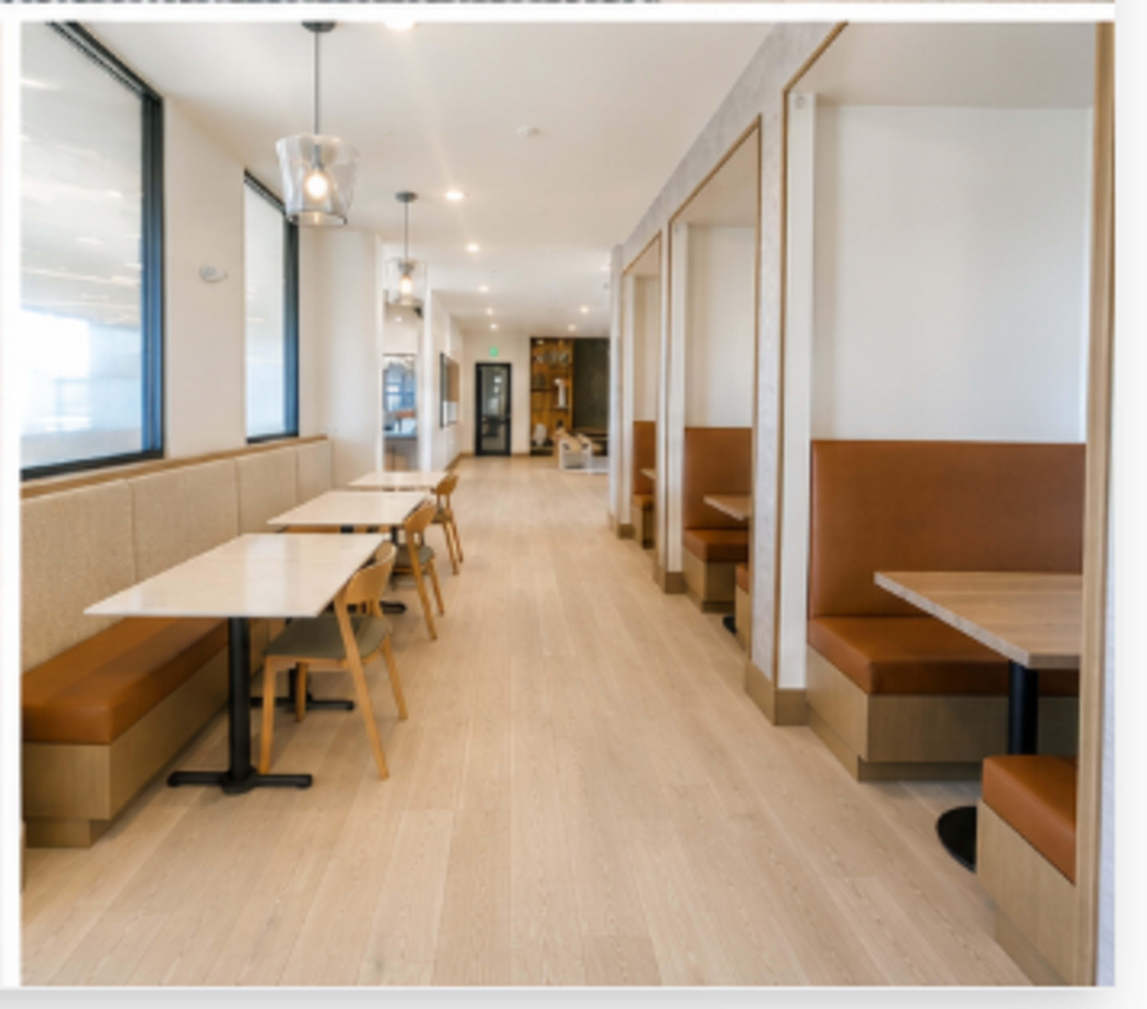 The image shows a modern restaurant interior featuring a long, narrow space with wooden tables and seating booths along the walls, illuminated by pendant lighting.