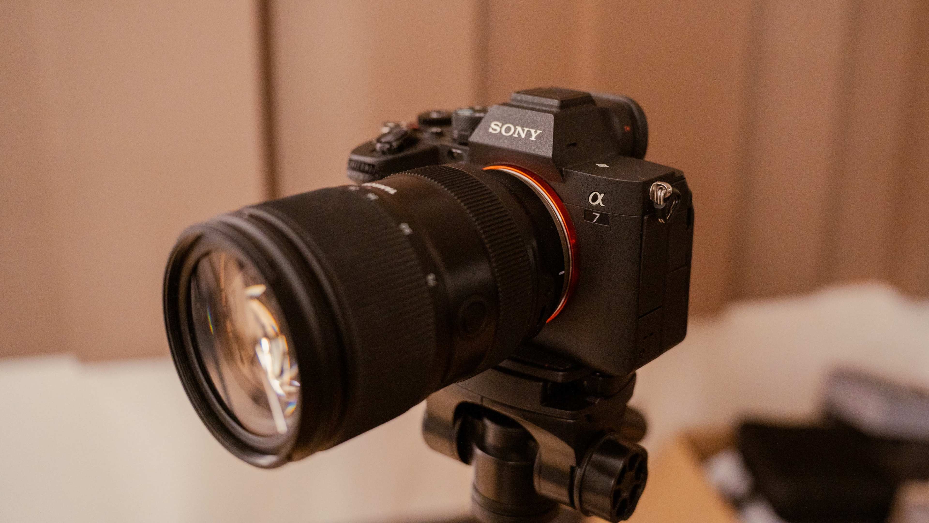 A Sony Alpha 7 camera with a large lens is mounted on a tripod in a room with brown curtains.