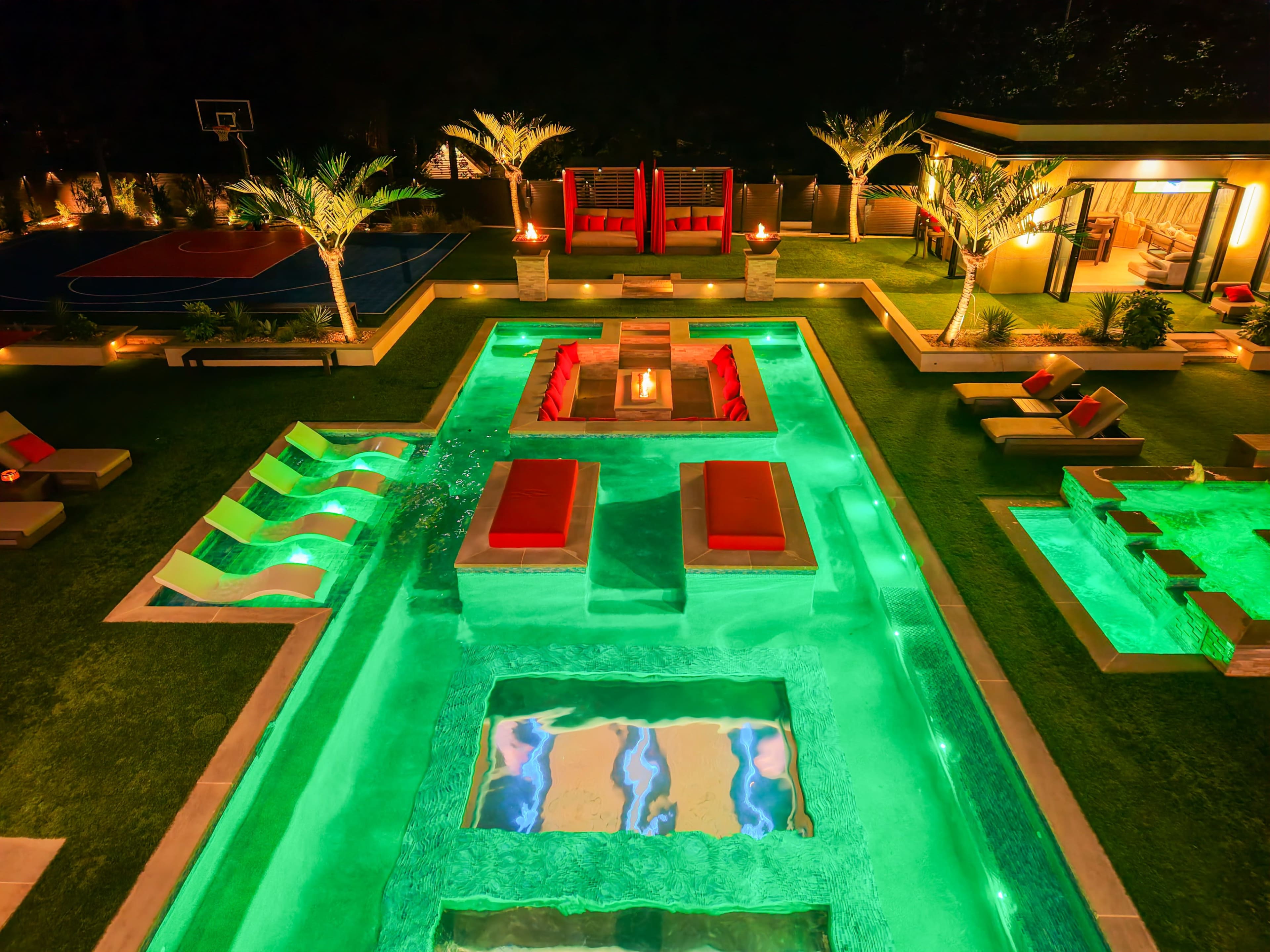 The image shows a luxurious outdoor pool area with illuminated water features, lounging spaces, and a basketball court in the background.