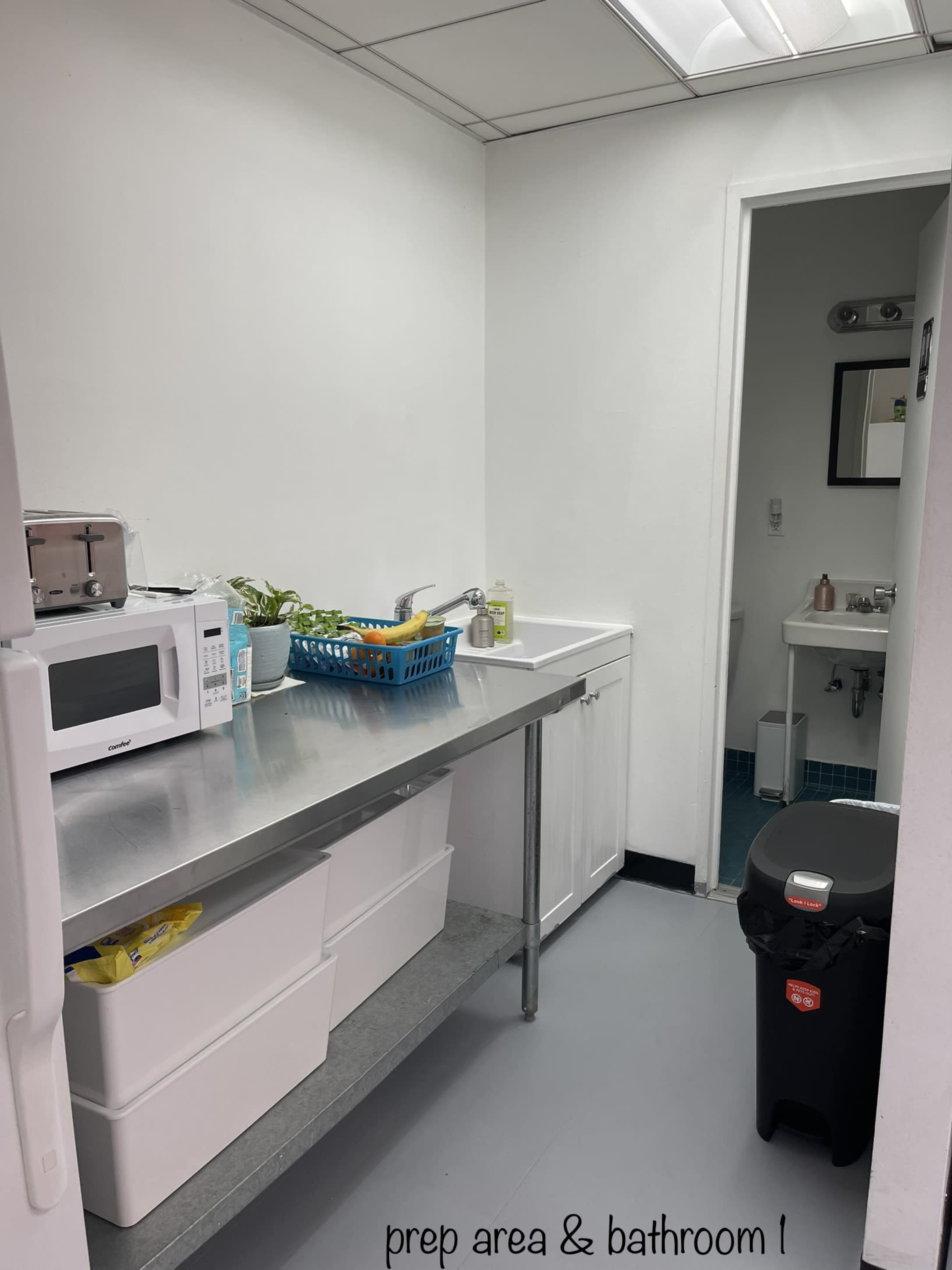 A kitchen prep area with a stainless steel table, a microwave, a small plant, and a trash can, alongside a bathroom visible through an open door.