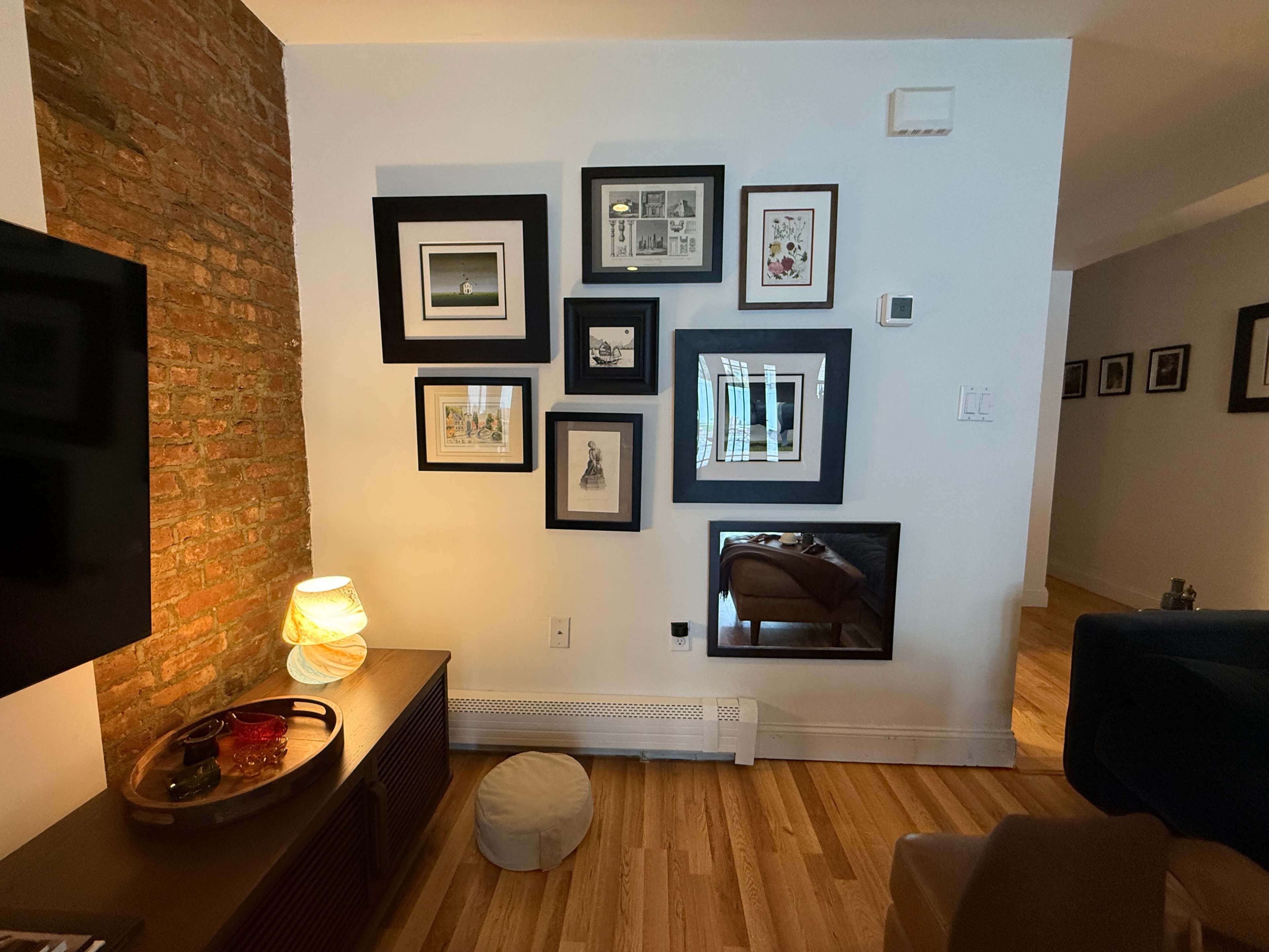 The scene features a living room corner with a brick wall displaying several framed pictures, a television on a wooden console, and a cozy seating area with a lamp.