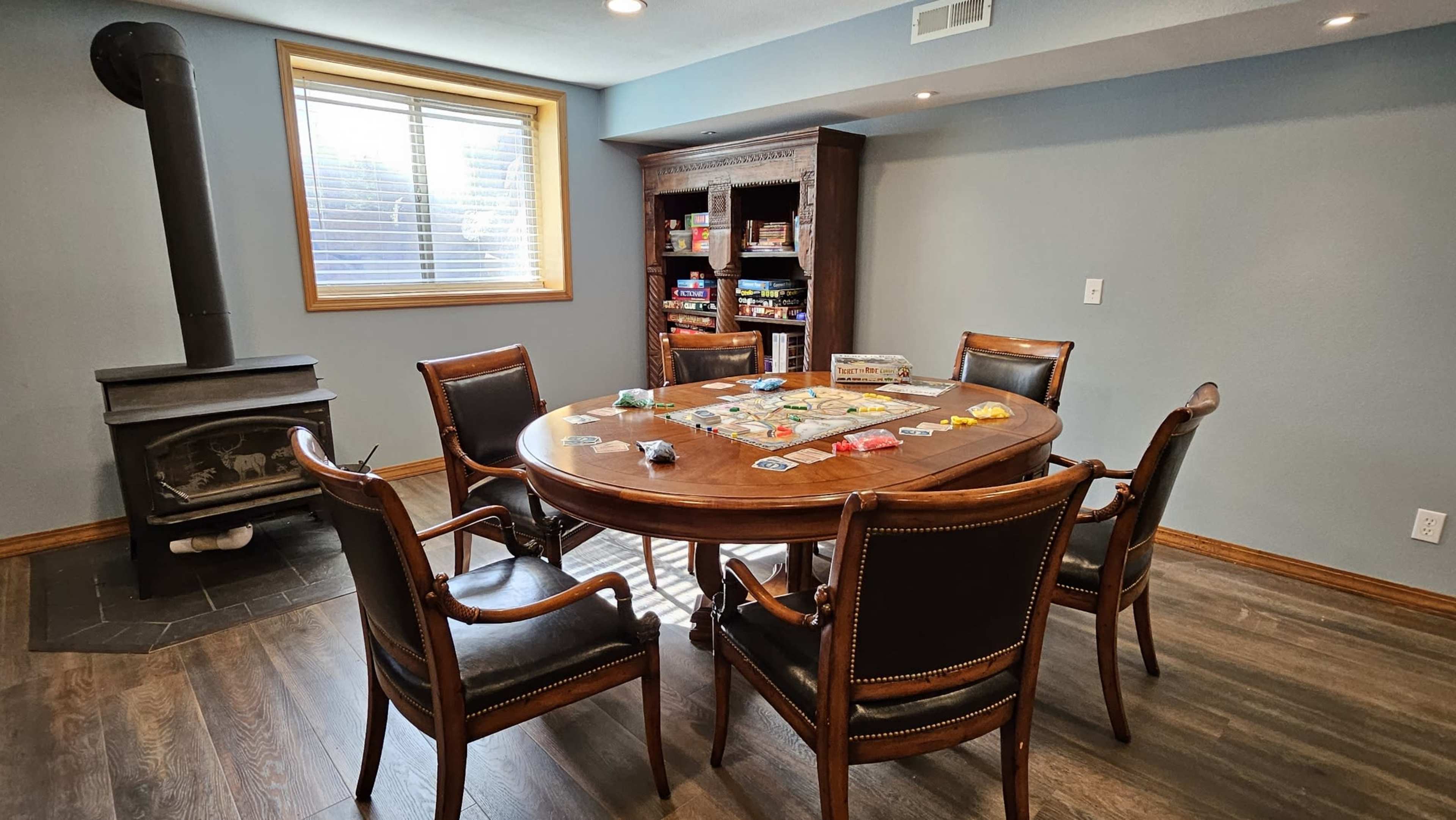A round wooden table surrounded by four chairs is set up for a board game in a well-lit room with a window and a wood stove.