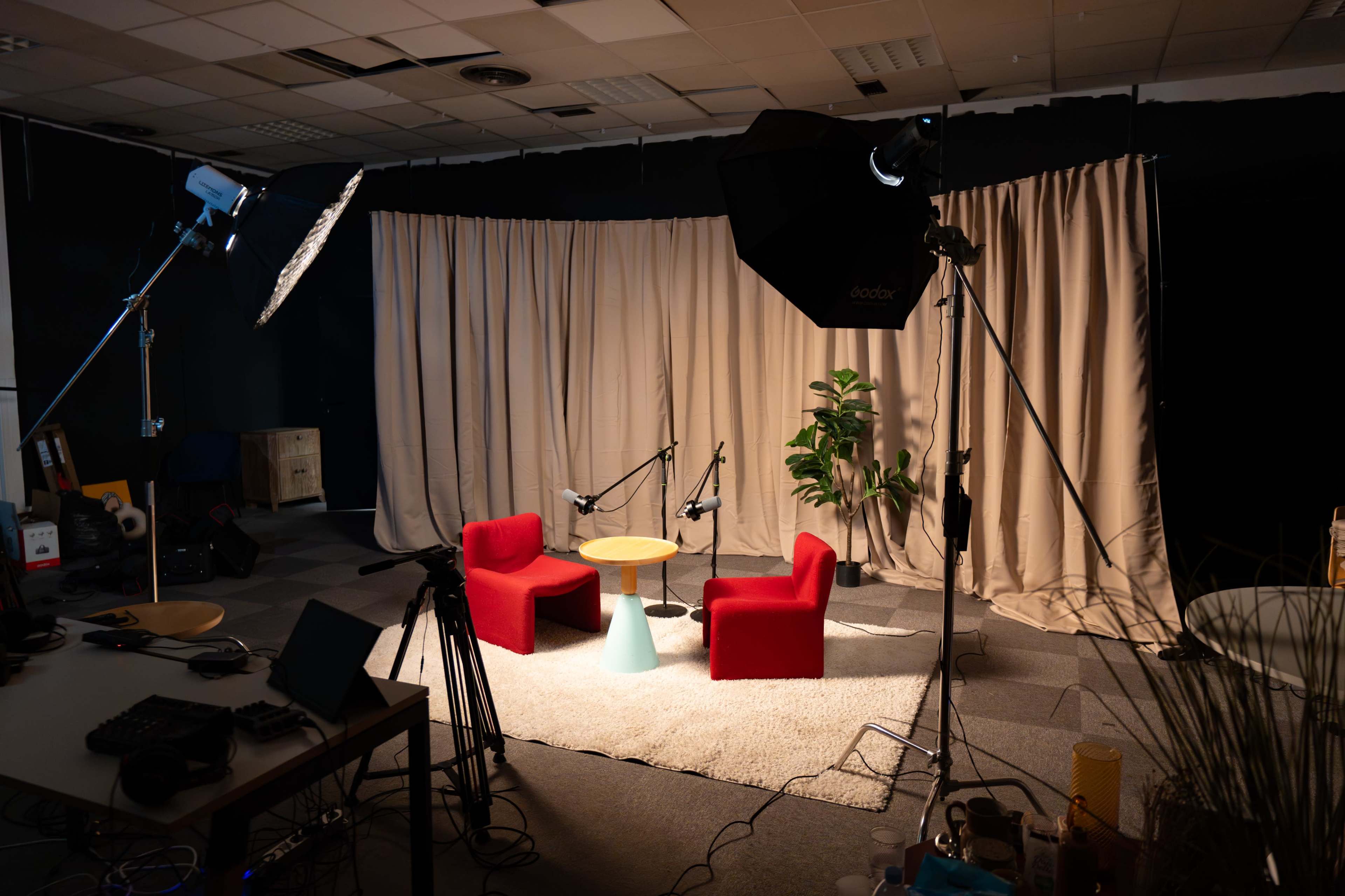 A small interview setup features two red chairs, a round table, and lighting equipment, surrounded by a soft rug and fabric backdrop.