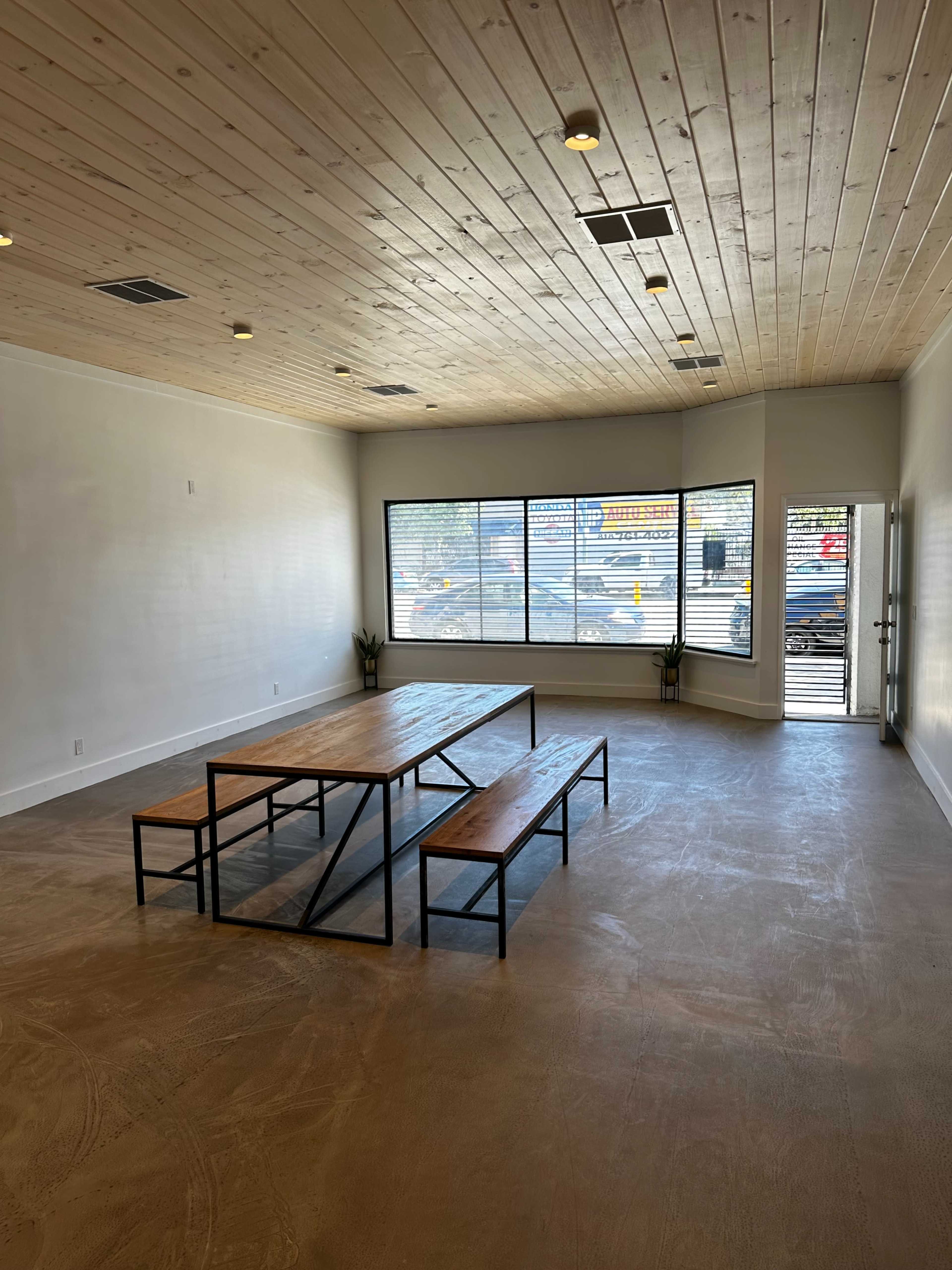 A spacious, well-lit interior featuring a wooden table with two benches and large windows allowing natural light to enter.