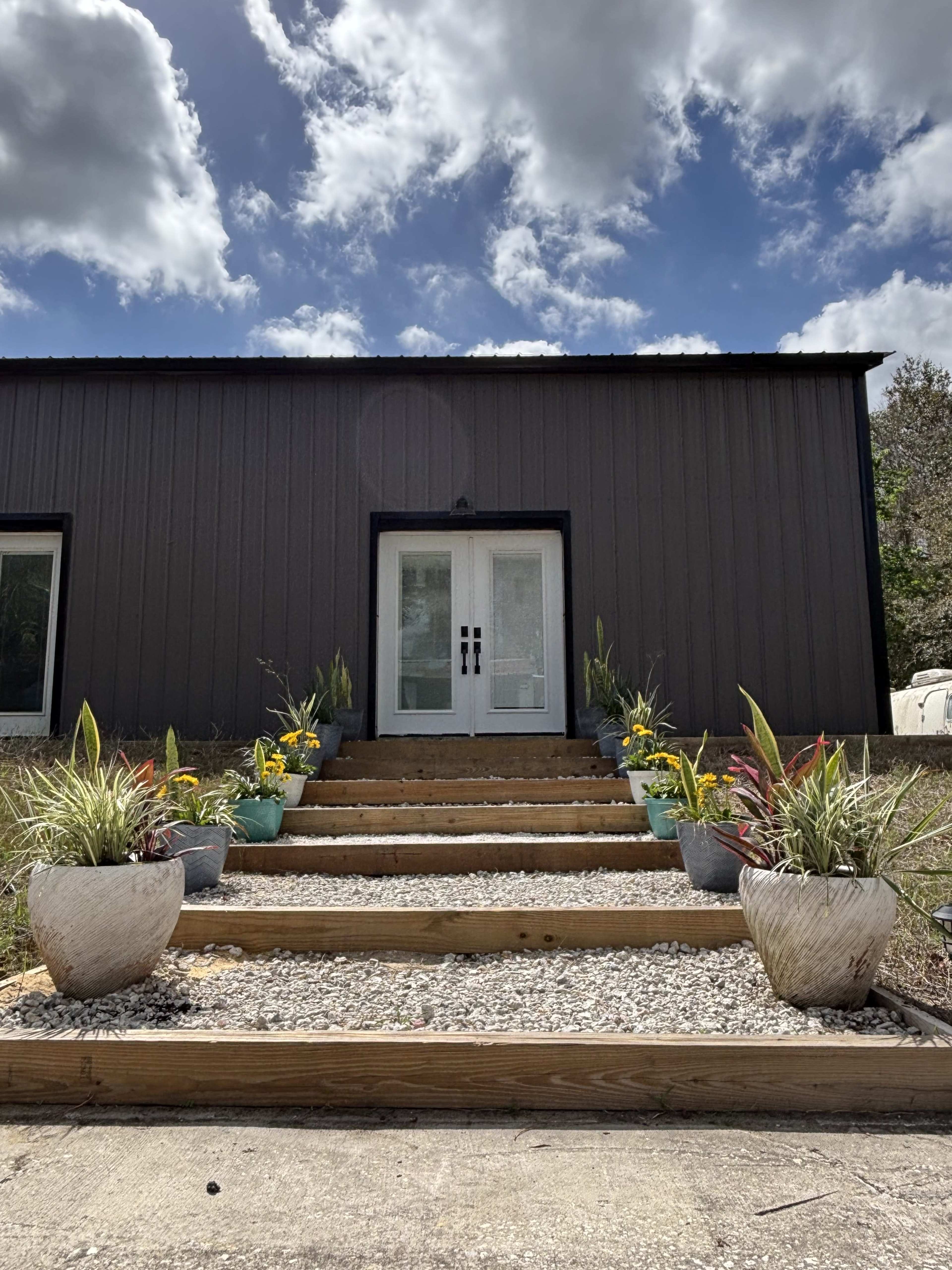 The image shows a staircase leading up to a modern black building with double doors, flanked by potted plants on either side.