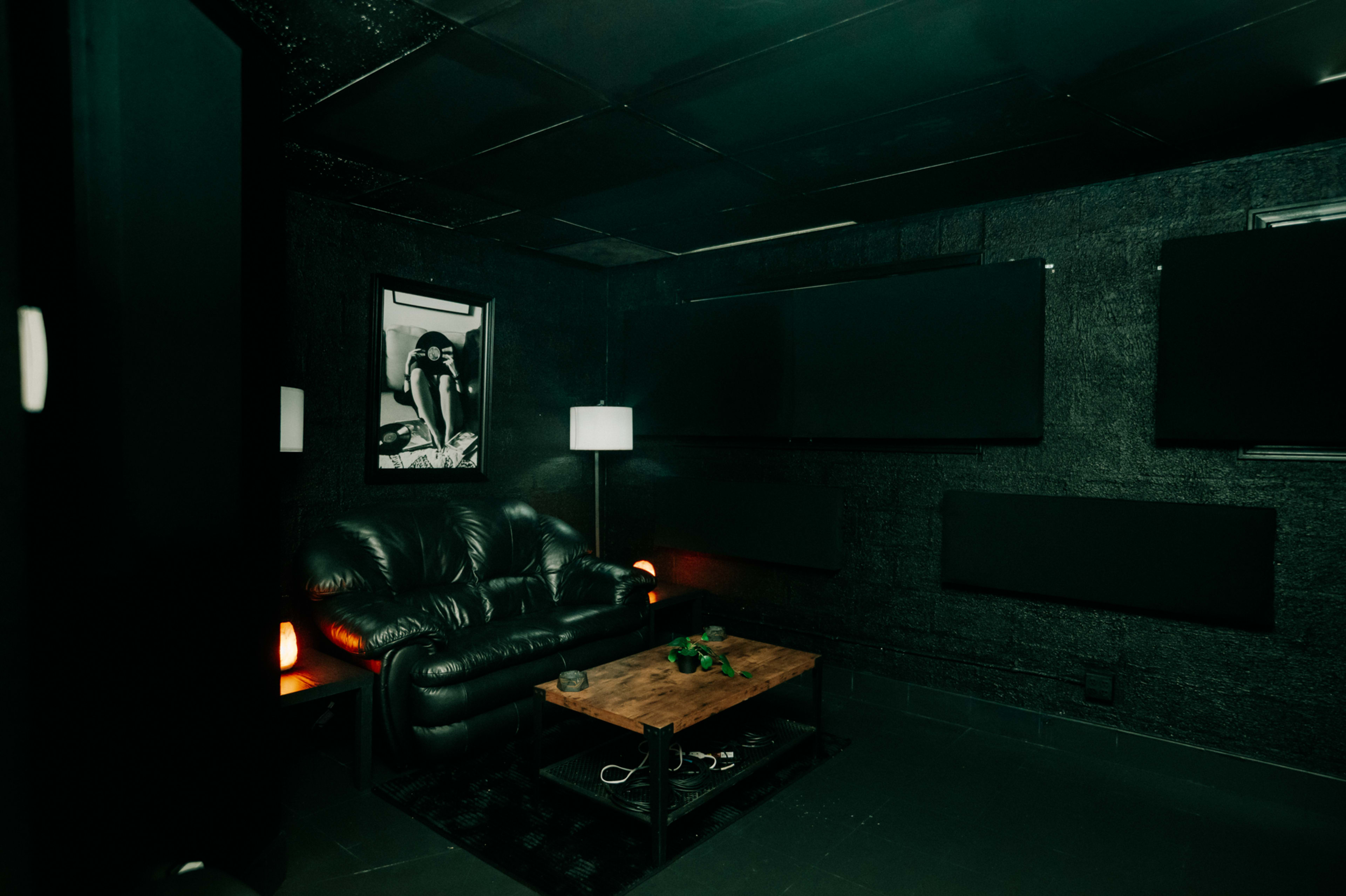 The image shows a darkened room featuring a black leather couch, a wooden coffee table, and several large black screens on the walls, illuminated by a lamp and two glowing salt lamps.