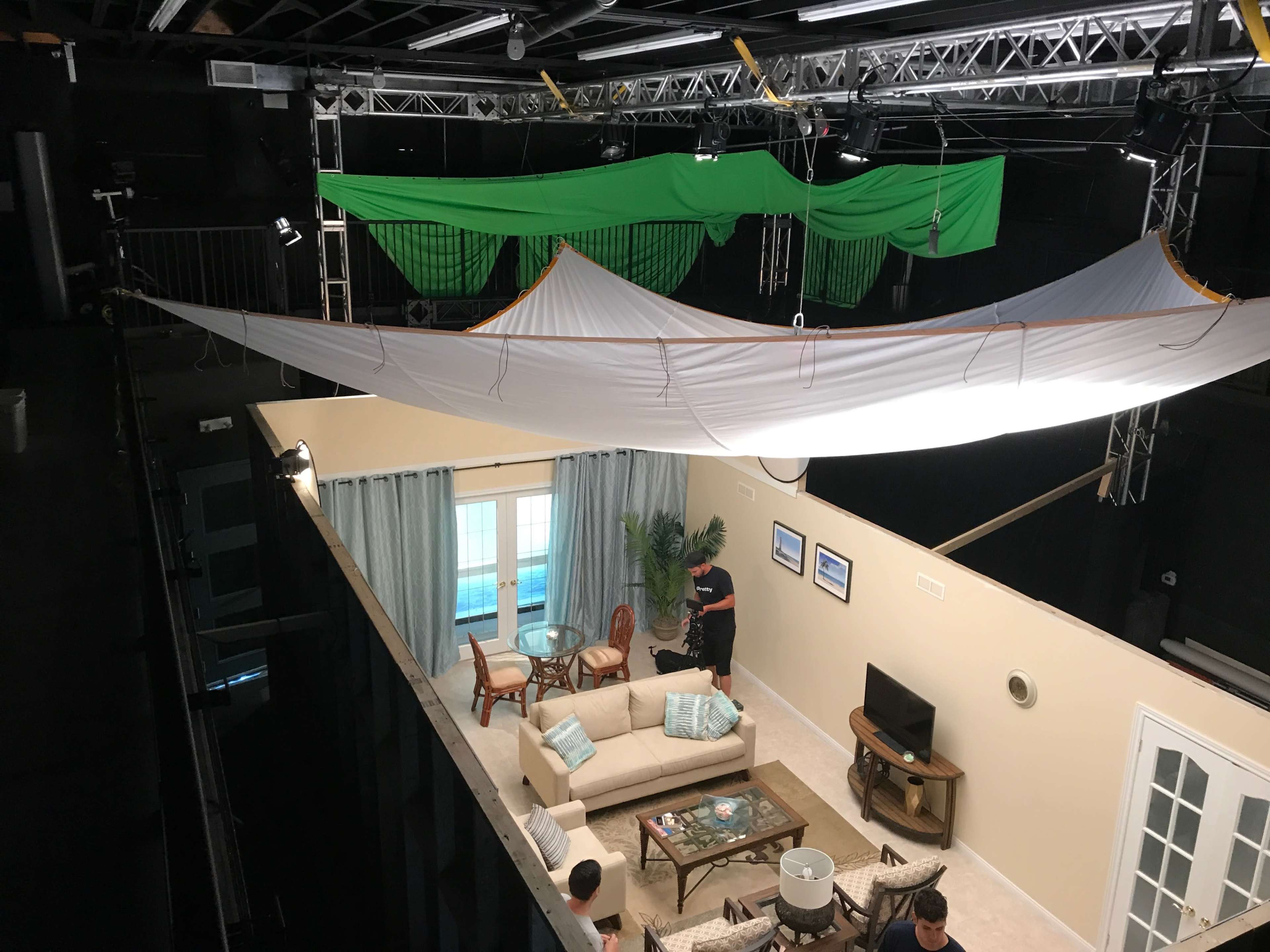 A spacious film set features a living room area with a sofa, chairs, and a television, while green screens are suspended above.