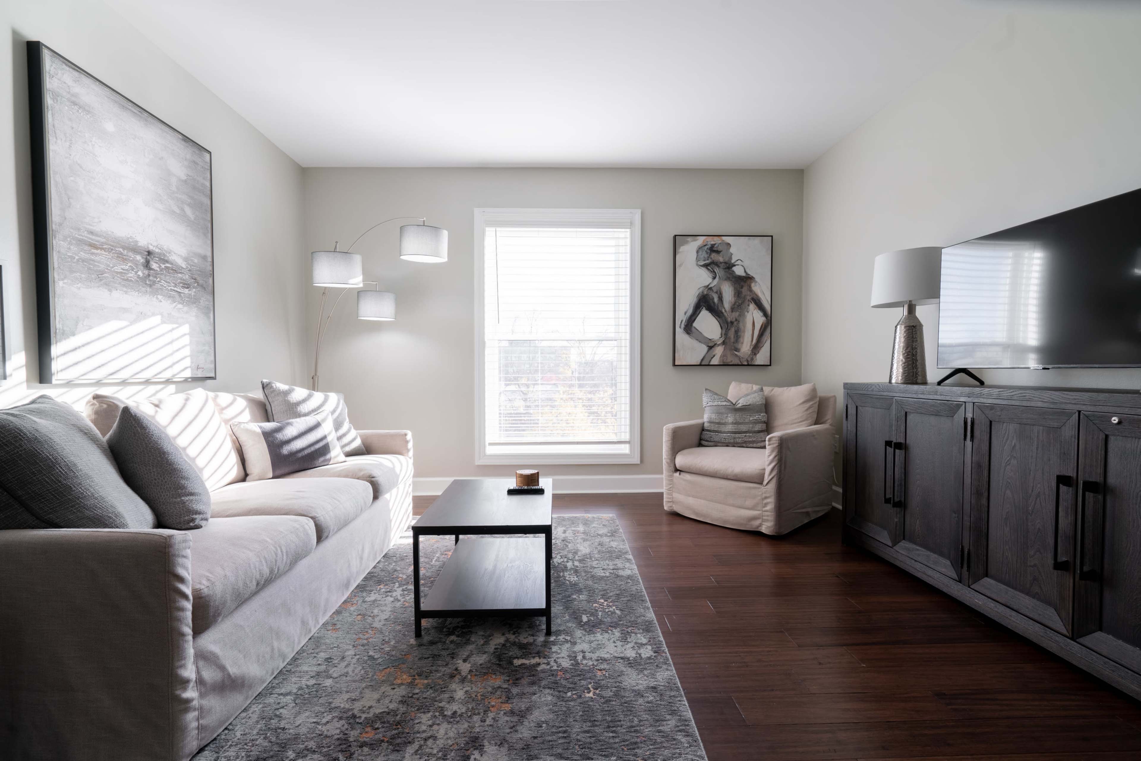 The image shows a modern living room with a light-colored sofa, a coffee table, a chair, and art on the walls, illuminated by natural light through a window.