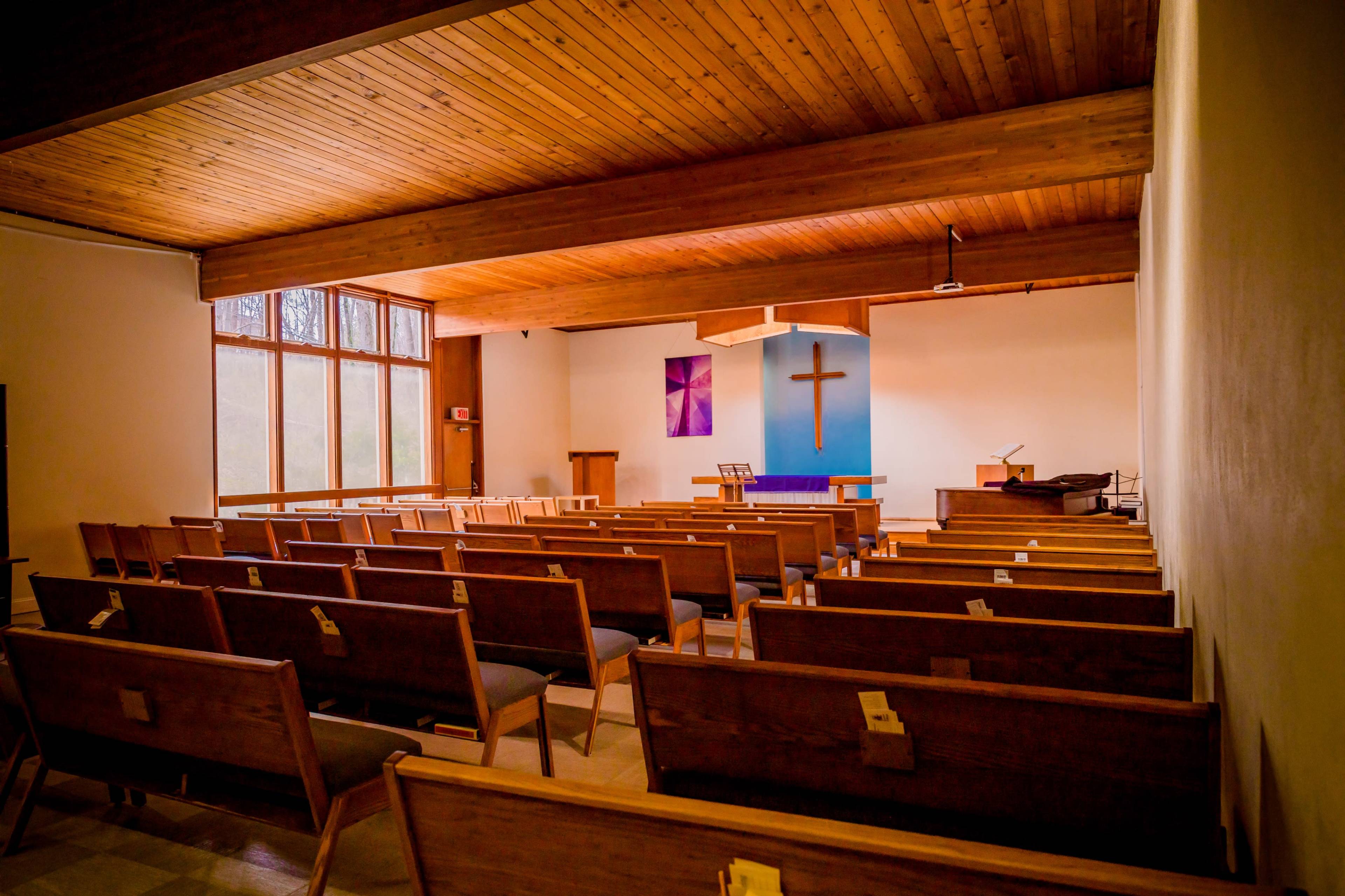 A spacious church interior with wooden pews arranged towards a front pulpit featuring a cross on the wall.